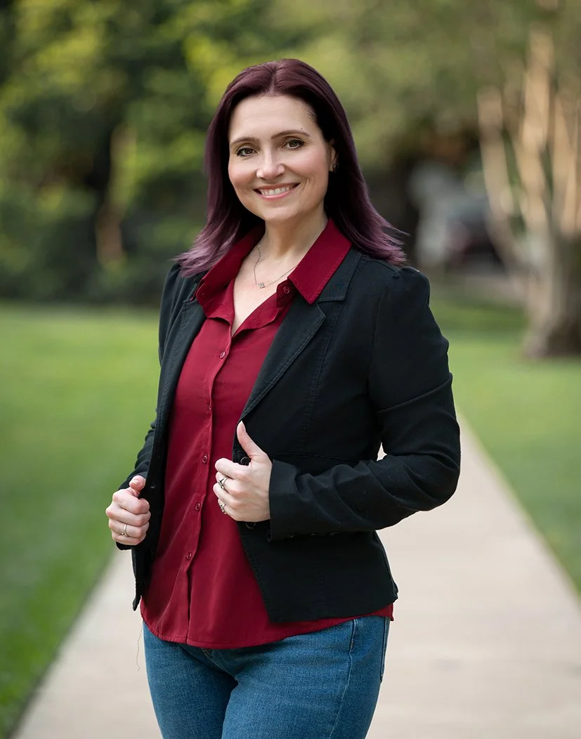A woman photographer with shoulder-length dark red hair smiling outdoors, wearing a black blazer over a red shirt and blue jeans, standing on a sidewalk in a park with green trees and grass in the background.