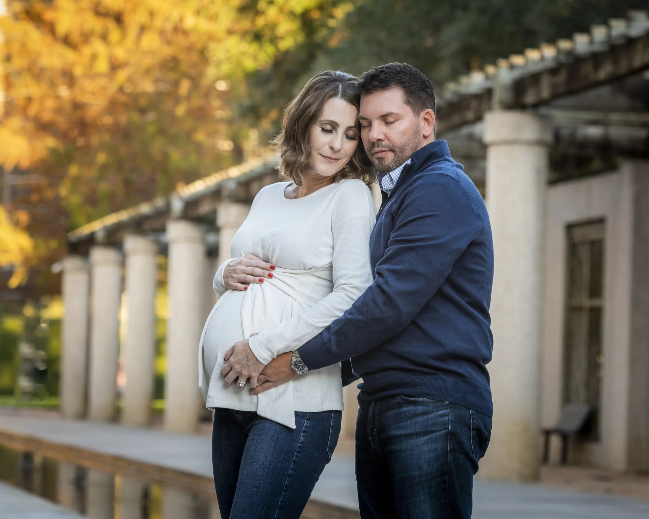 A pregnant woman and a man are standing close together outdoors with their eyes closed, gently touching the woman's belly, surrounded by trees with fall foliage.