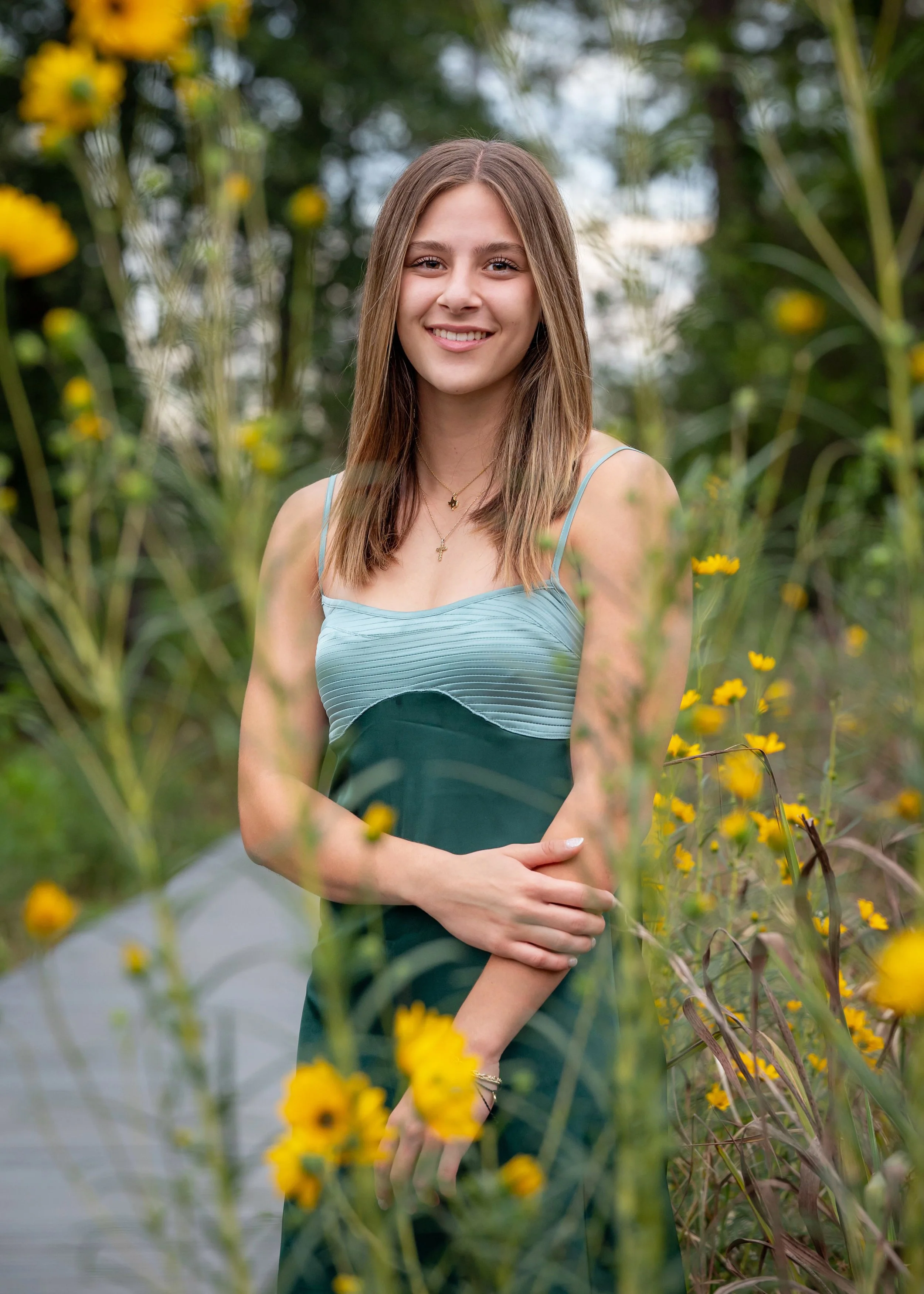 A senior girl with brown hair smiling, standing outdoors among yellow flowers and greenery, wearing a light blue sleeveless dress and jewelry.