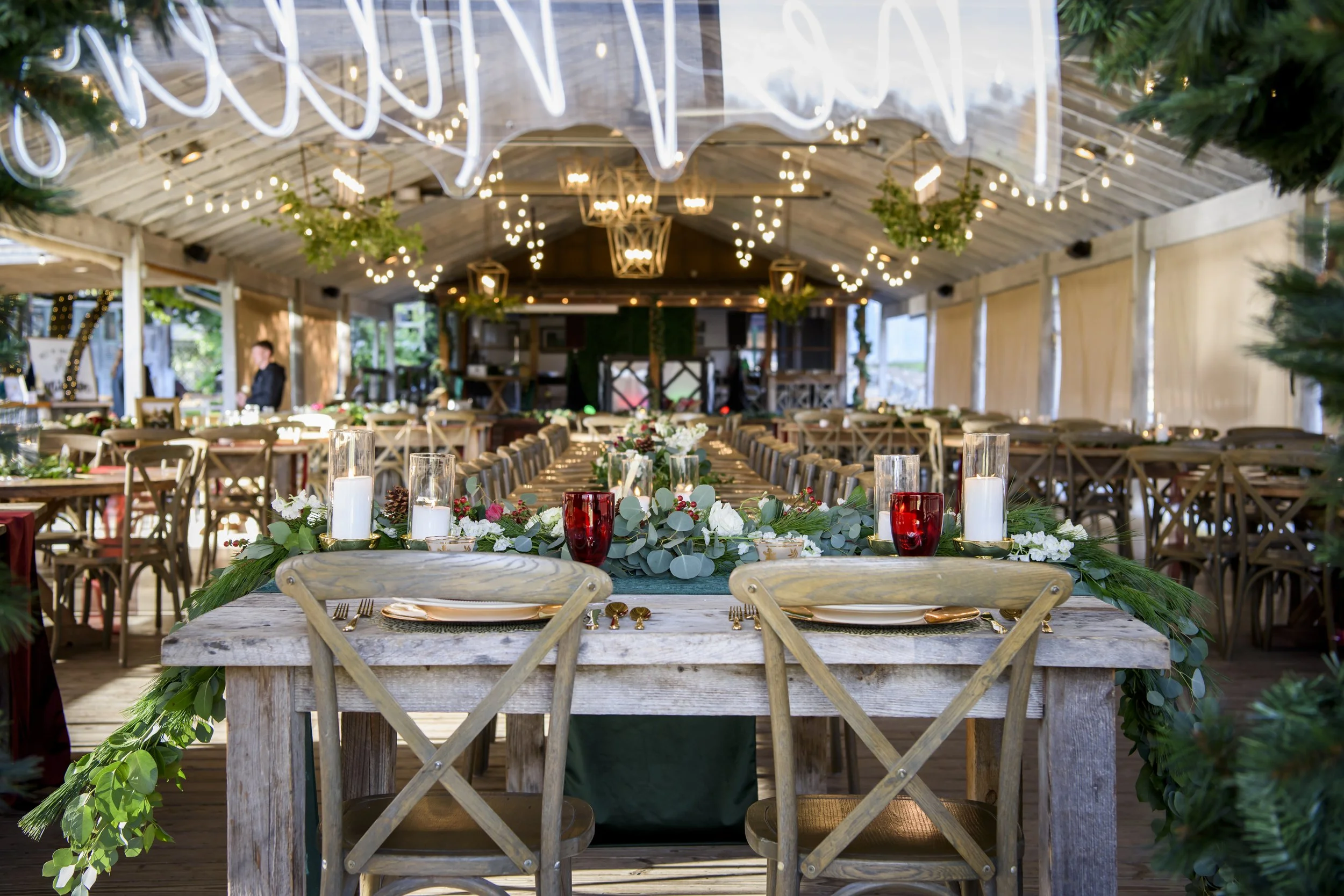 Indoor wedding reception decorated with string lights, candles, and floral arrangements on a long wooden table, with additional tables and greenery in the background.