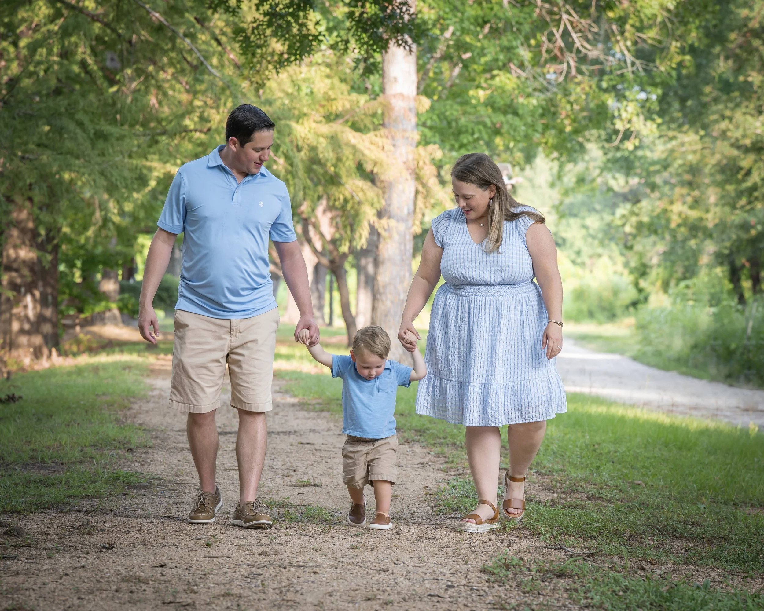 A man, woman, and little boy walking outdoors on a dirt path lined with grass, surrounded by green trees, in a park or forest.