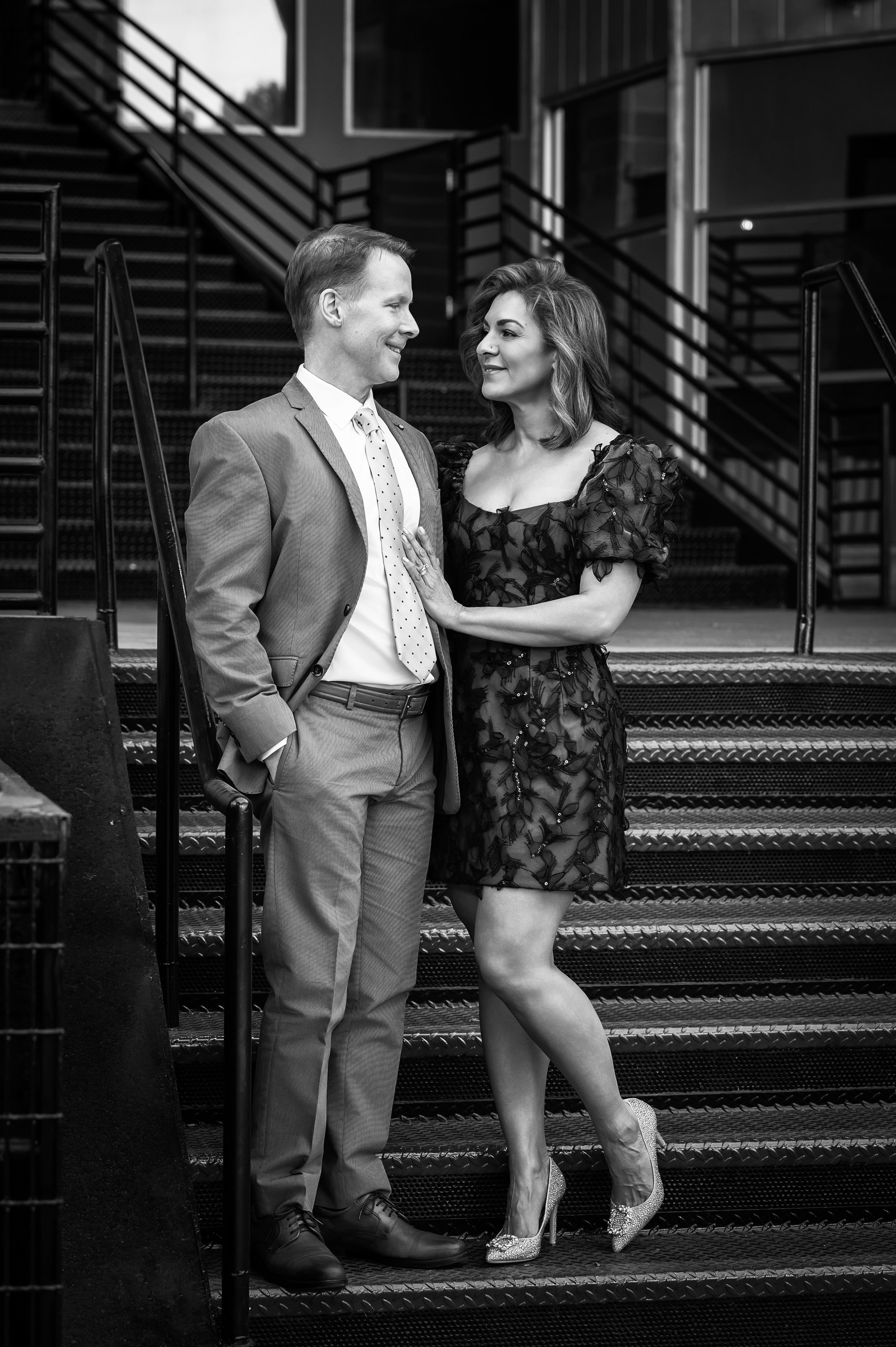 Black and white photo of a man and woman standing on a metal staircase outside a building. The man is dressed in a suit and tie, smiling at the woman. The woman is wearing a short, lacy dress with floral details and high heels, gazing at the man.