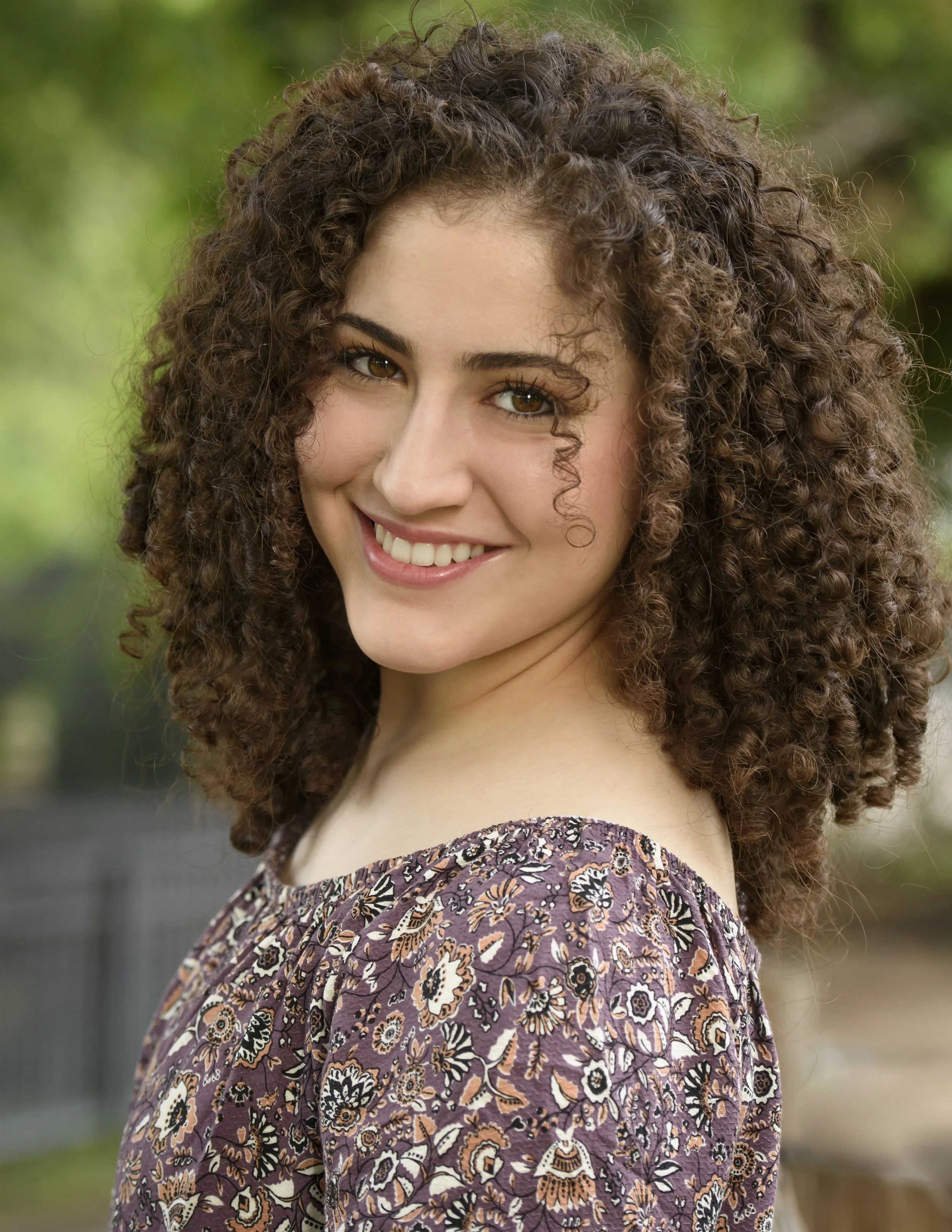 A young woman with curly brown hair, hazel eyes, and a warm smile, wearing a floral-patterned purple dress, standing outdoors with blurred green foliage in the background.