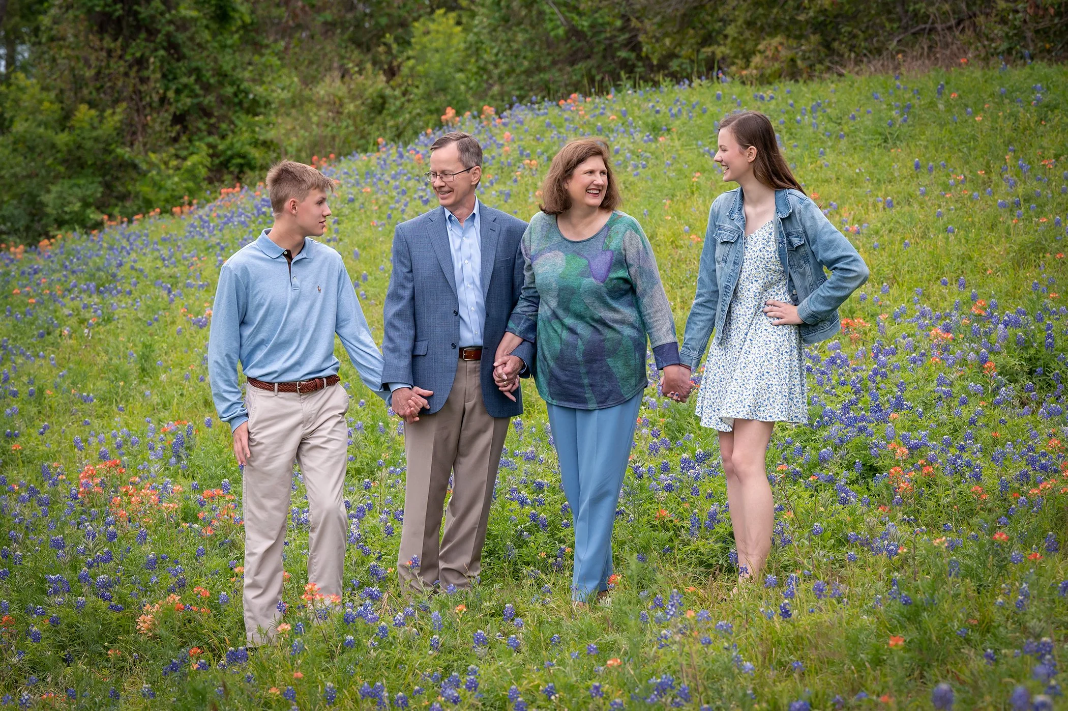 A family of four, including a teenage boy, a man, a woman, and a teenage girl, holding hands and walking through a field of wildflowers and bluebonnets with trees in the background, smiling and enjoying each other's company.