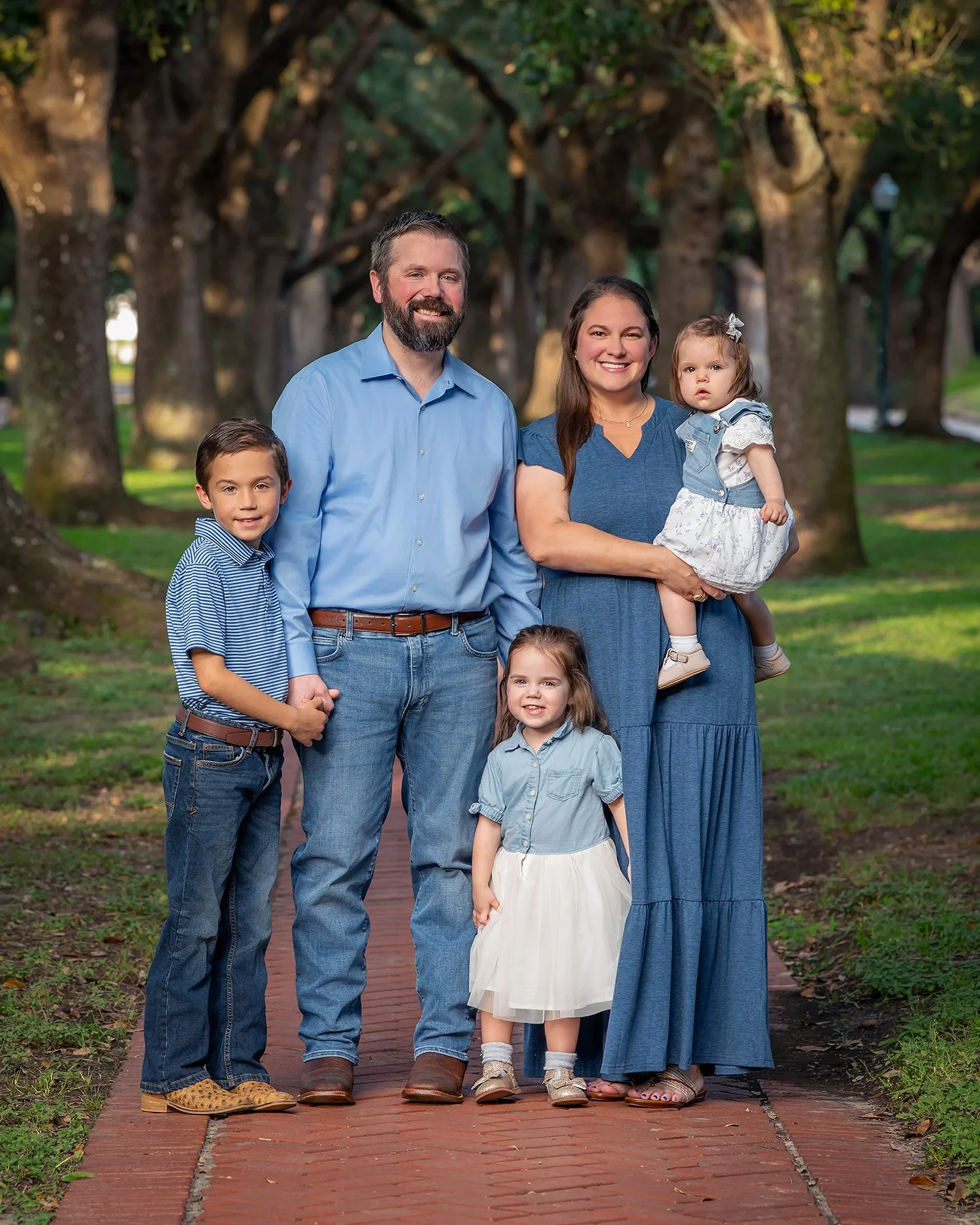 Family of six posing on a brick path in a park with large trees in the background. The family includes two adult men, one adult woman, and three young children, all smiling and wearing casual clothing.