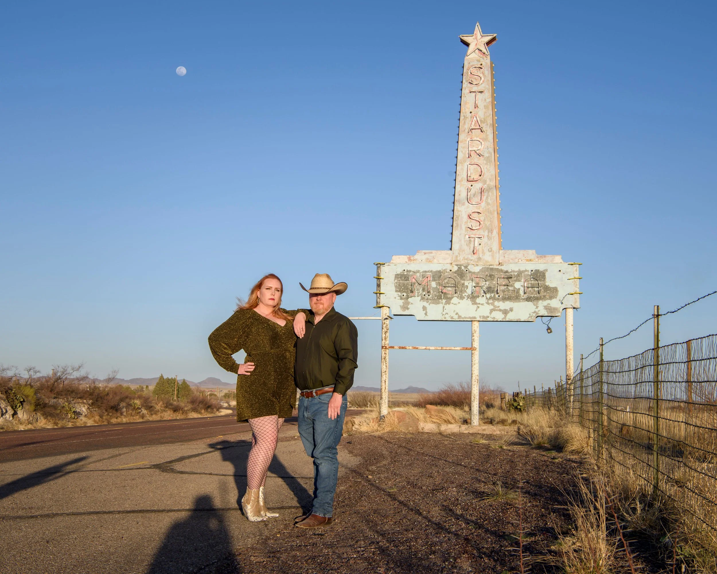 A woman and a man standing near a vintage neon sign on a deserted road in a desert landscape with a clear blue sky and the moon visible in the background.
