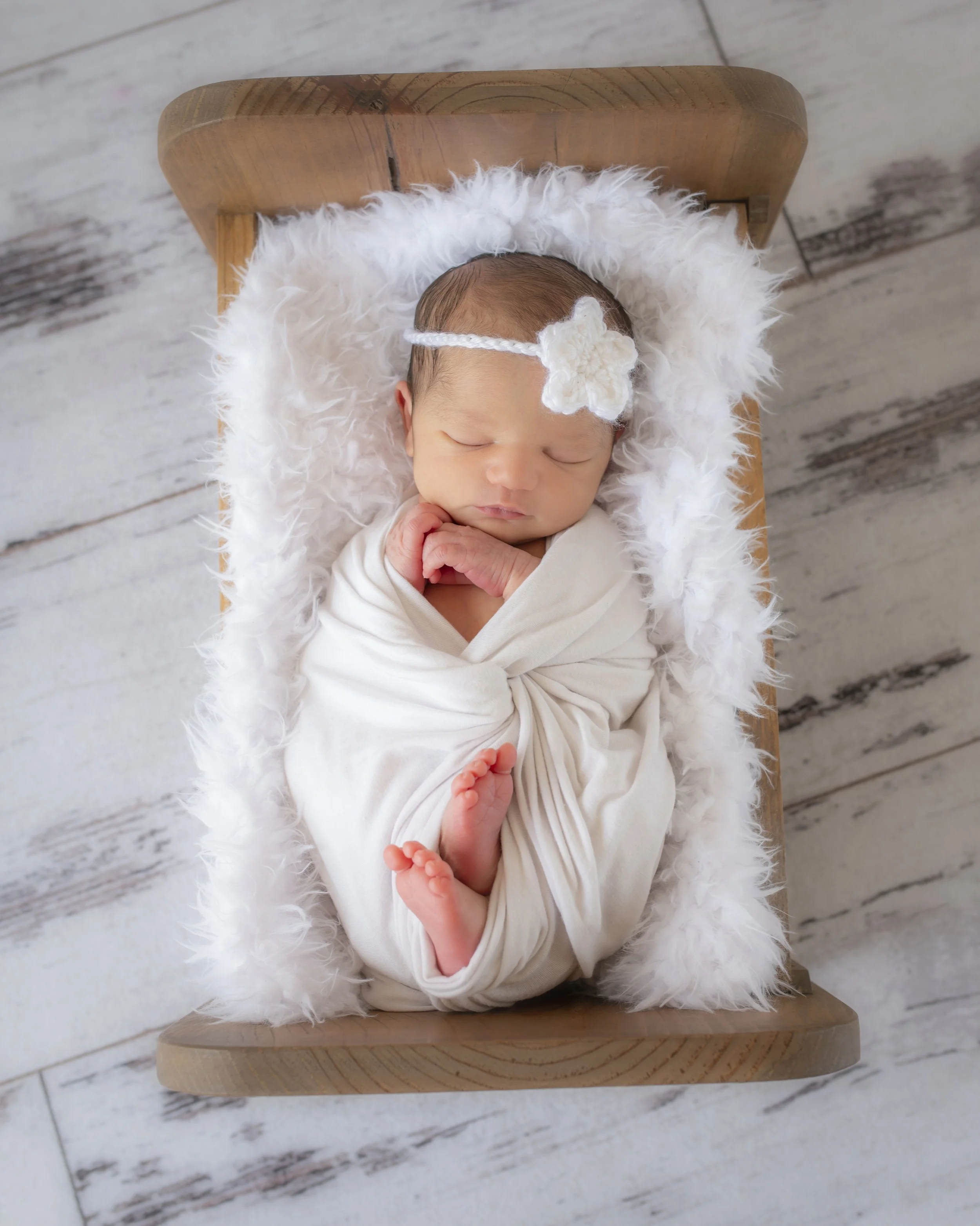 A newborn baby sleeping on a fluffy white blanket on a wooden chair, decorated with a white flower headband.
