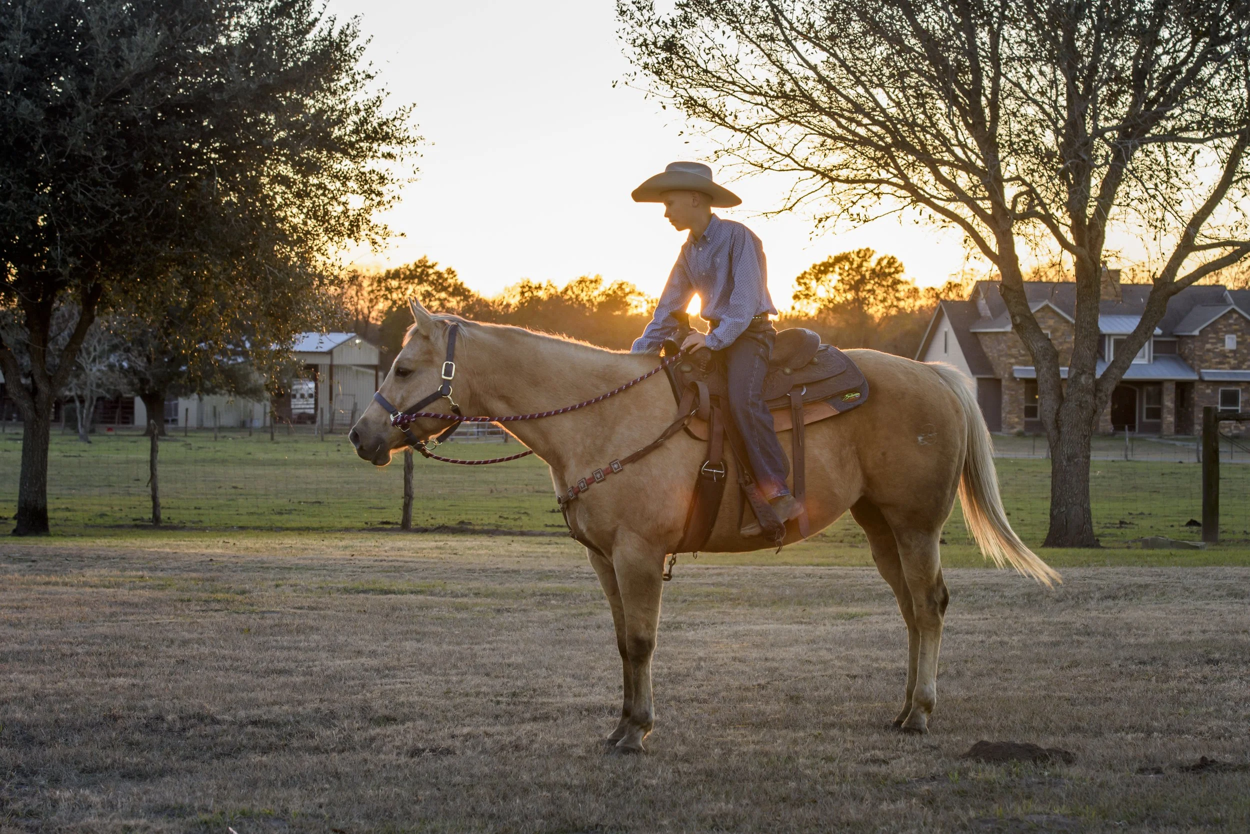 A teenager boy in cowboy attire riding a light-colored horse in a grassy field during sunset, with trees and residential buildings in the background.