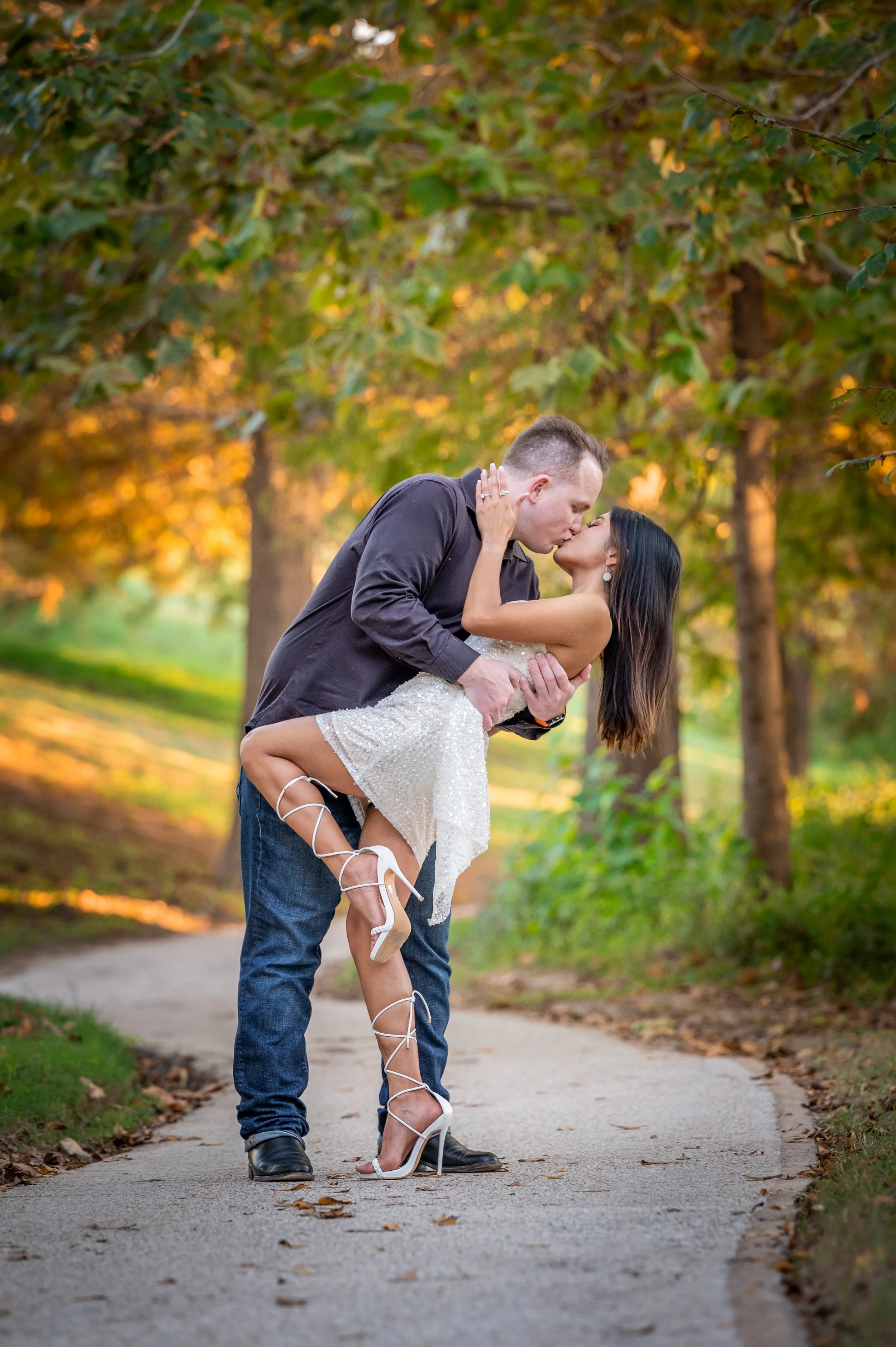 A man in dark clothing is holding a woman in a white dress, dipping her while kissing outdoors on a pathway surrounded by trees with green and orange leaves.