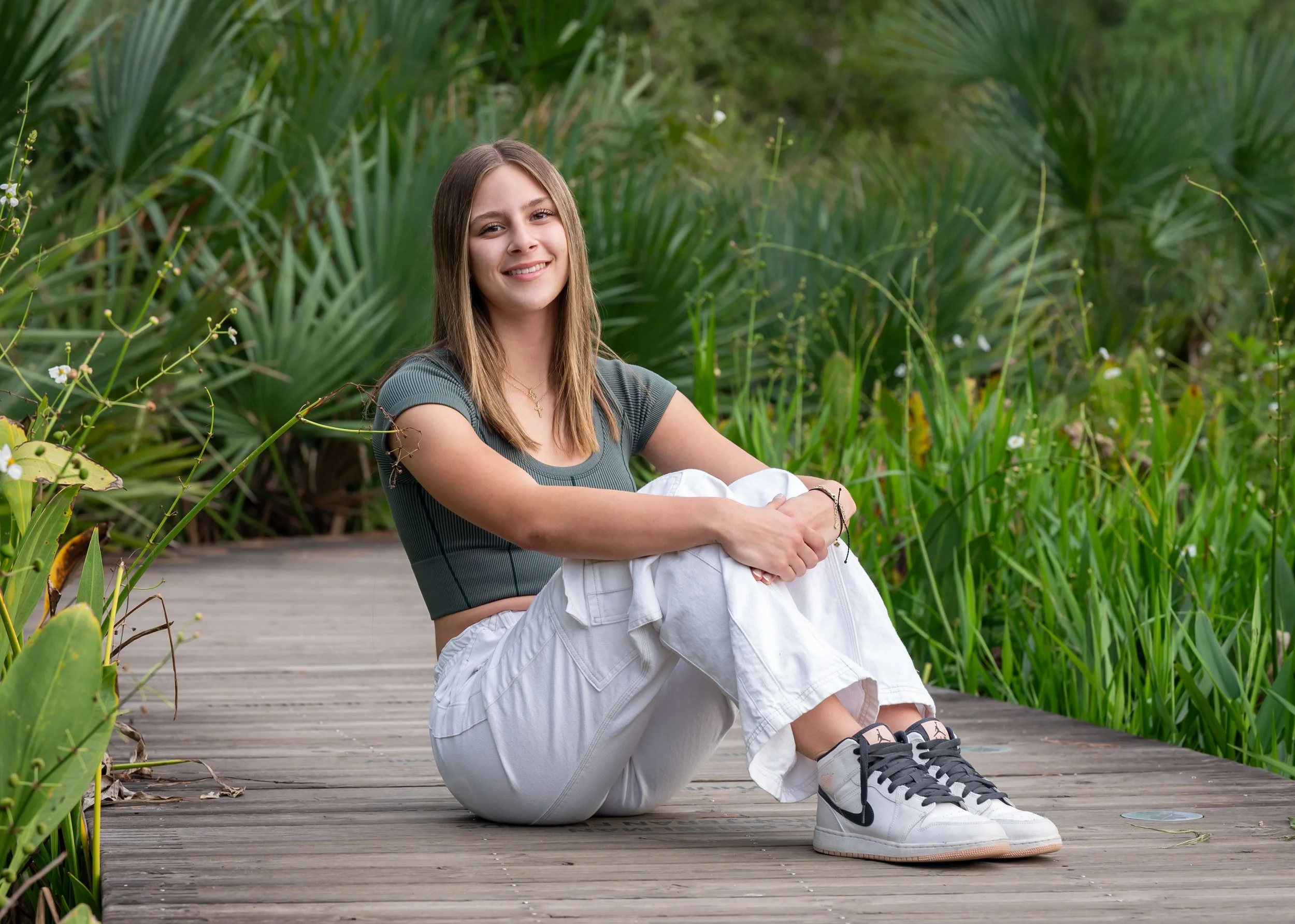 A senior girl with long light brown hair sitting on a wooden path surrounded by green plants, smiling at the camera, wearing a gray t-shirt, white pants, and sneakers.