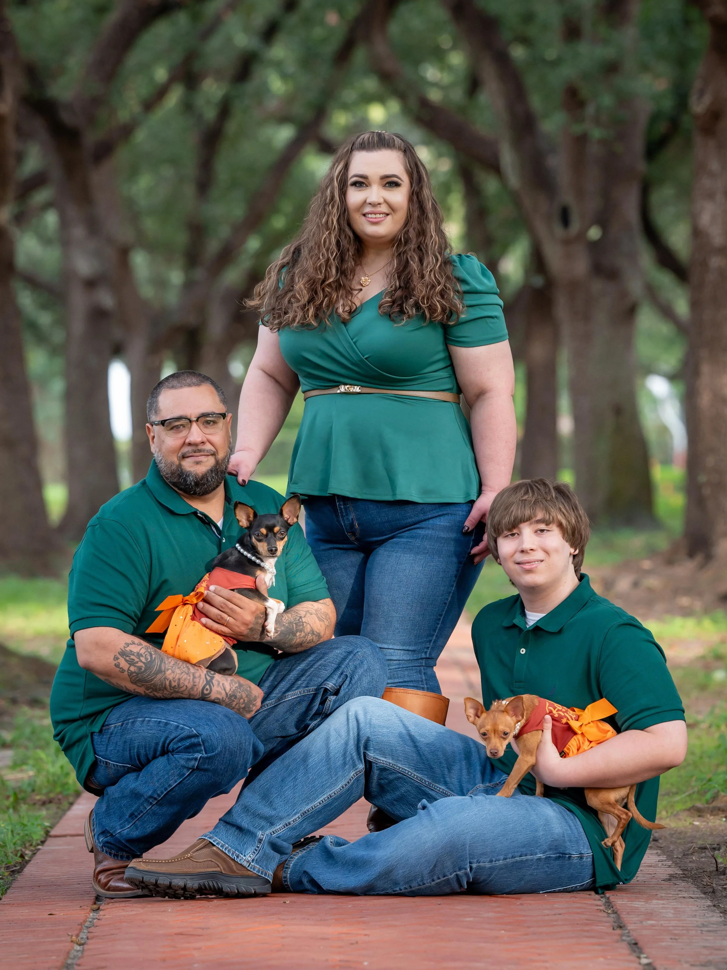 A diverse group of three people and two dogs in a park with large trees in the background. They are sitting and standing on a brick path and are dressed in green clothing, smiling at the camera.
