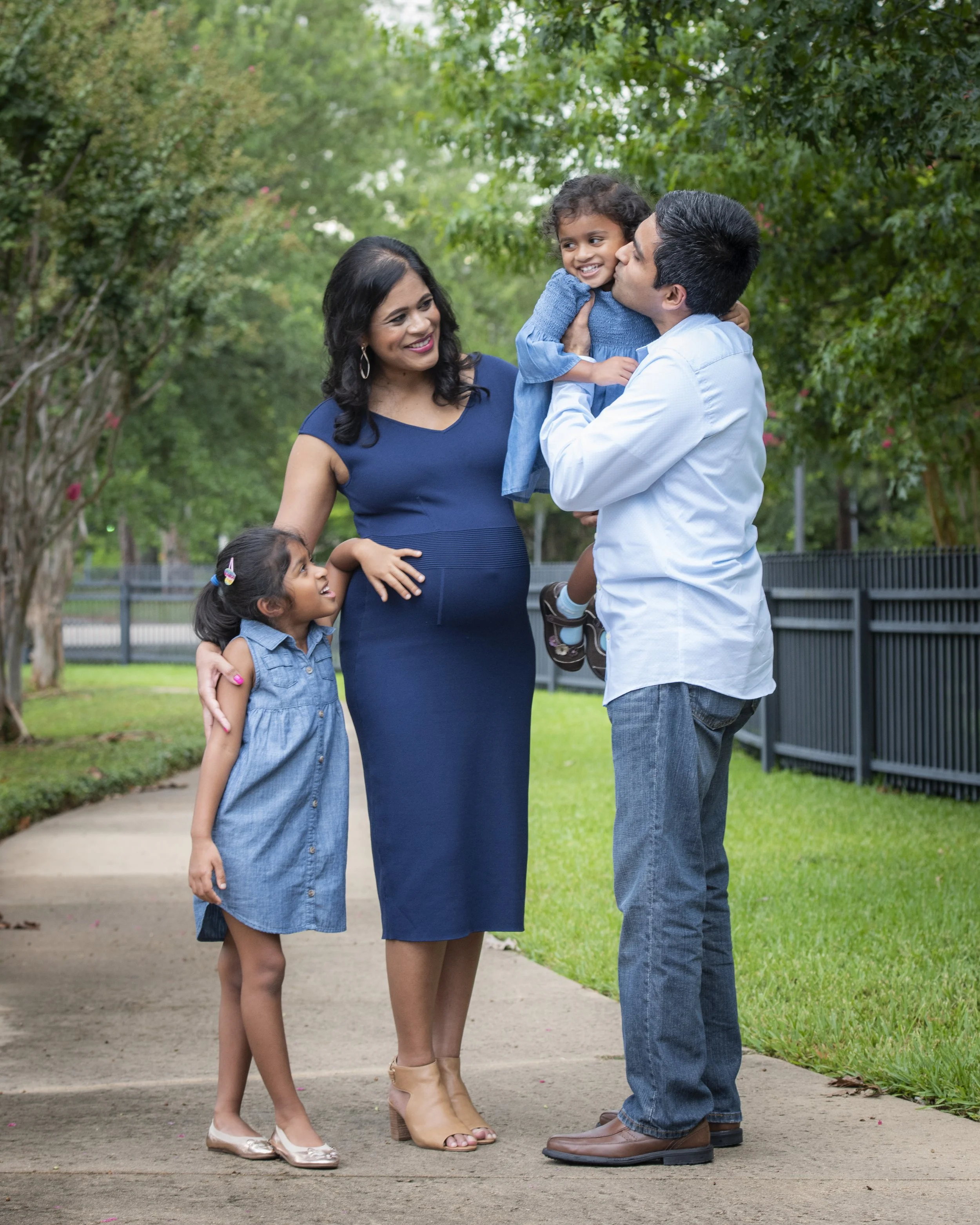 A family of four visits in a park with trees, including a pregnant woman in a blue dress, a man in a white shirt holding a smiling young girl, and a young girl in a denim dress.