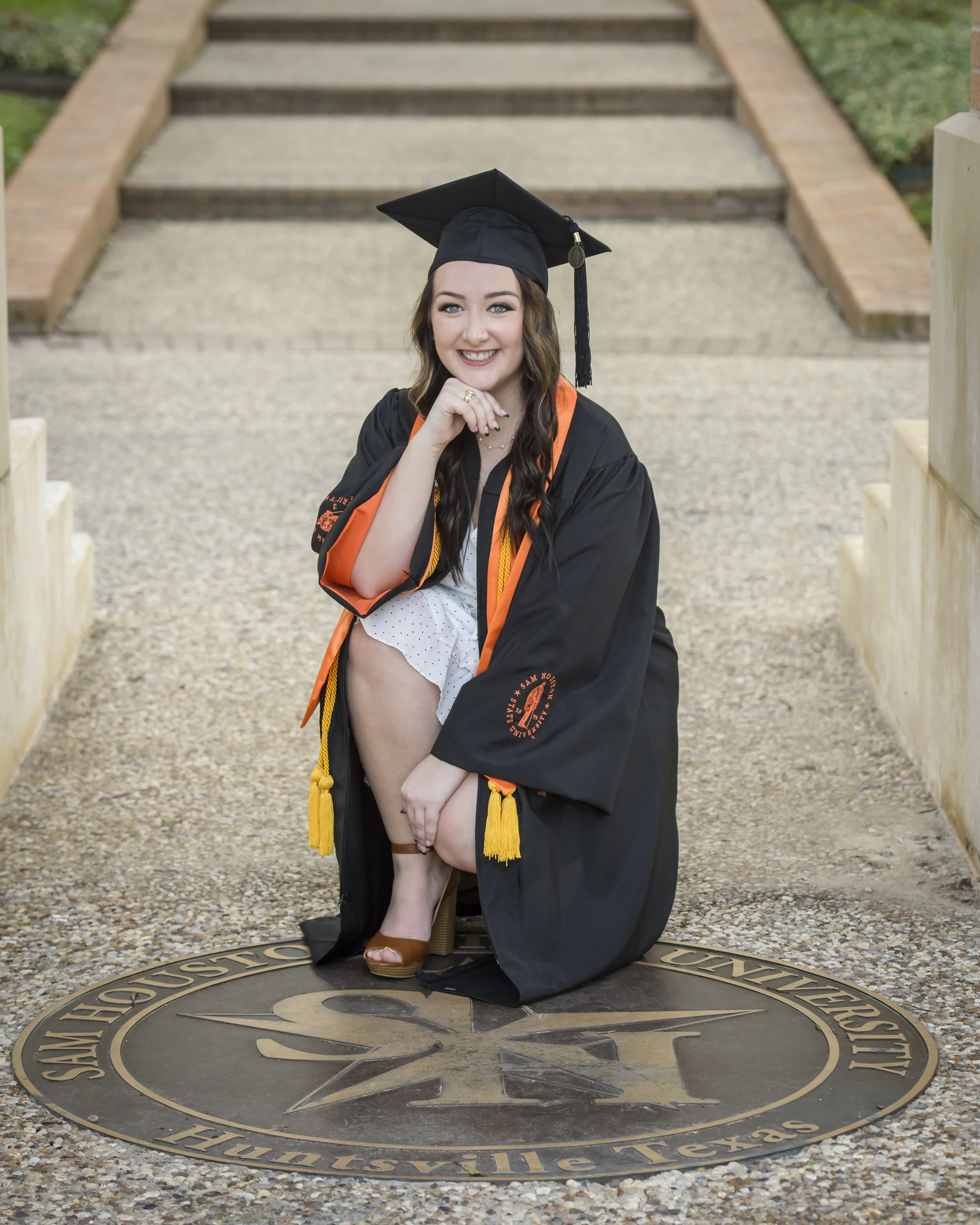 A young woman in a black graduation gown and cap is kneeling on a medallion engraved with university information, smiling with her hand resting on her chin. She is wearing orange and yellow honor cords, standing on a stone walkway with steps behind h