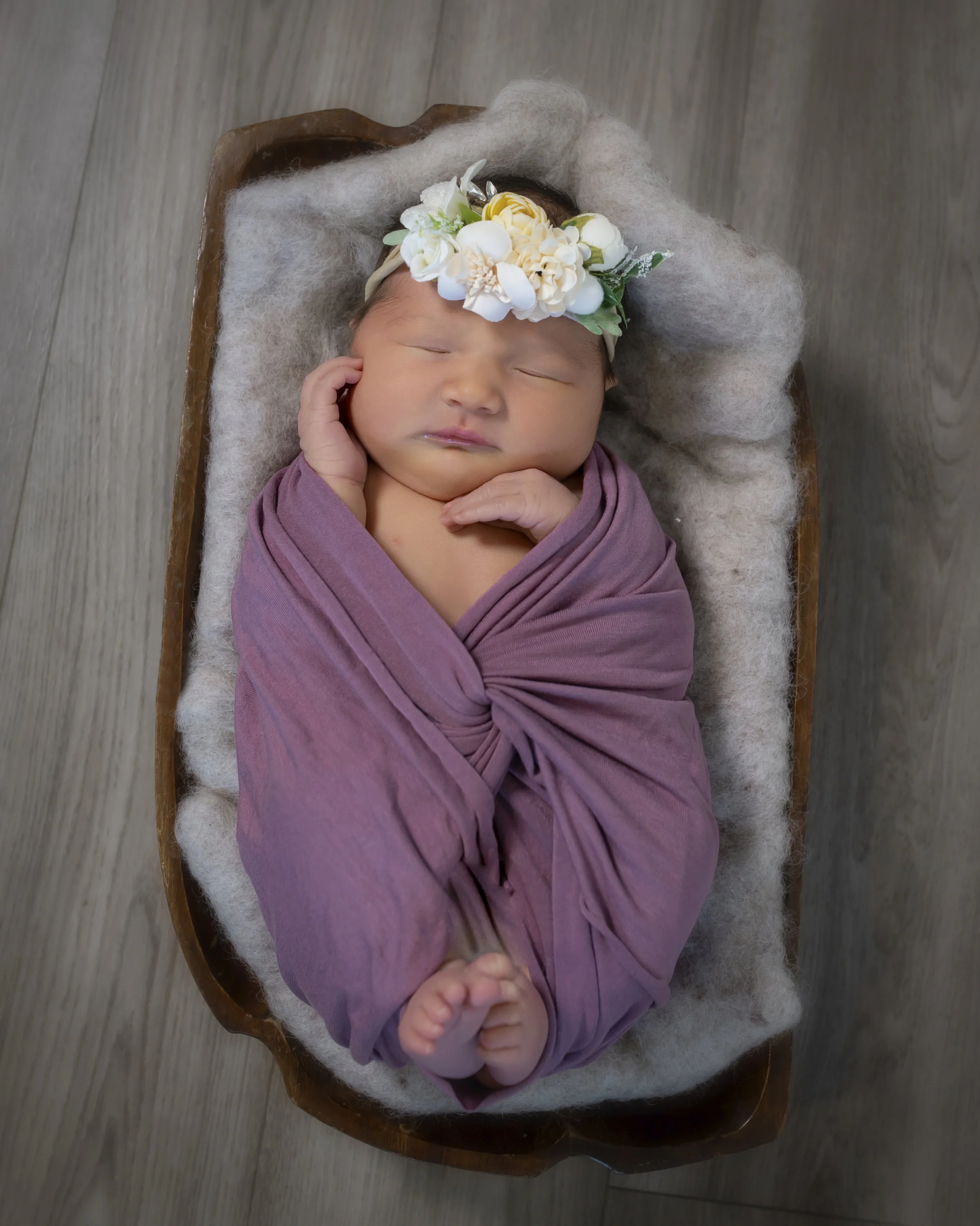 A sleeping newborn baby wrapped in a mauve blanket, wearing a floral headband with white flowers, lying in a small wooden bed with a soft beige blanket, placed on a wooden floor.