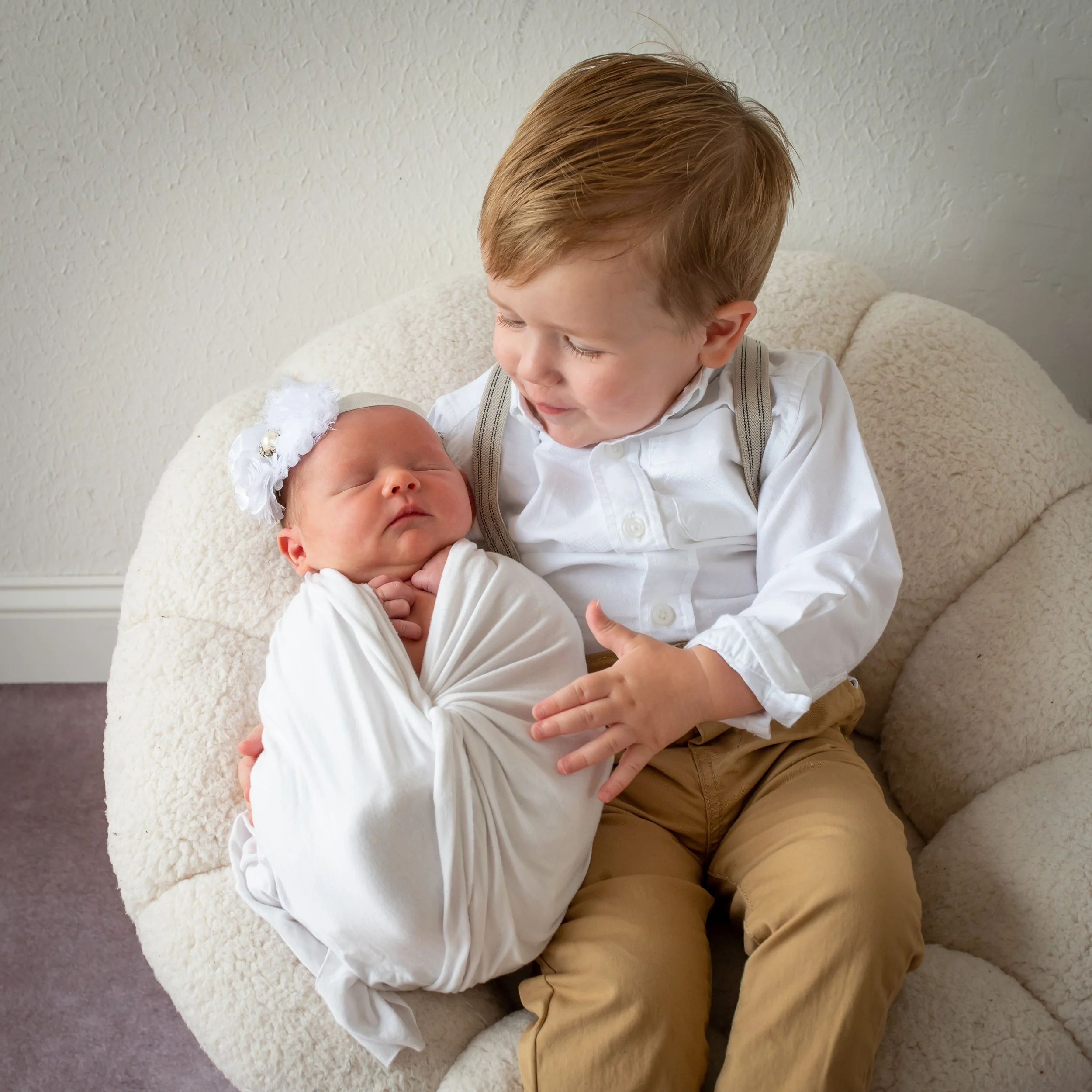 A young boy with red hair sitting on a fluffy ottoman, looking at and holding a newborn baby girl dressed in white with a floral headband.