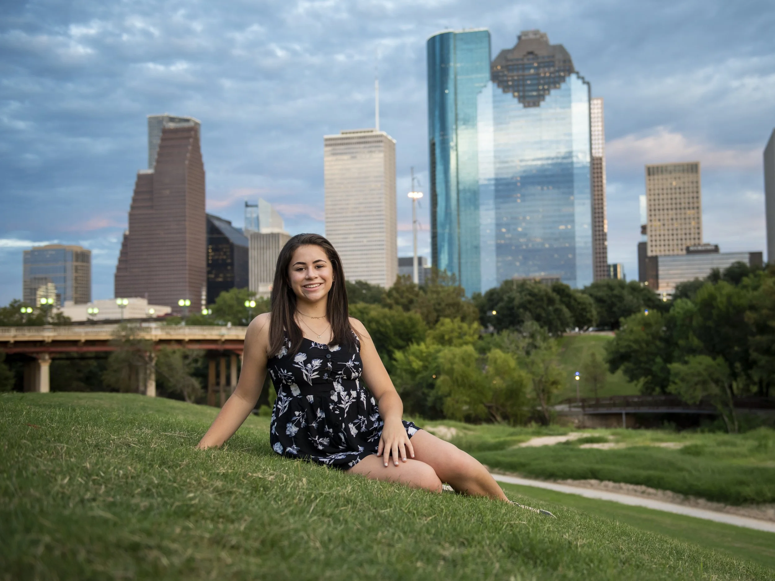 Senior girl sitting on grass in a park with a Houston skyline background featuring tall modern buildings, under a partly cloudy sky.