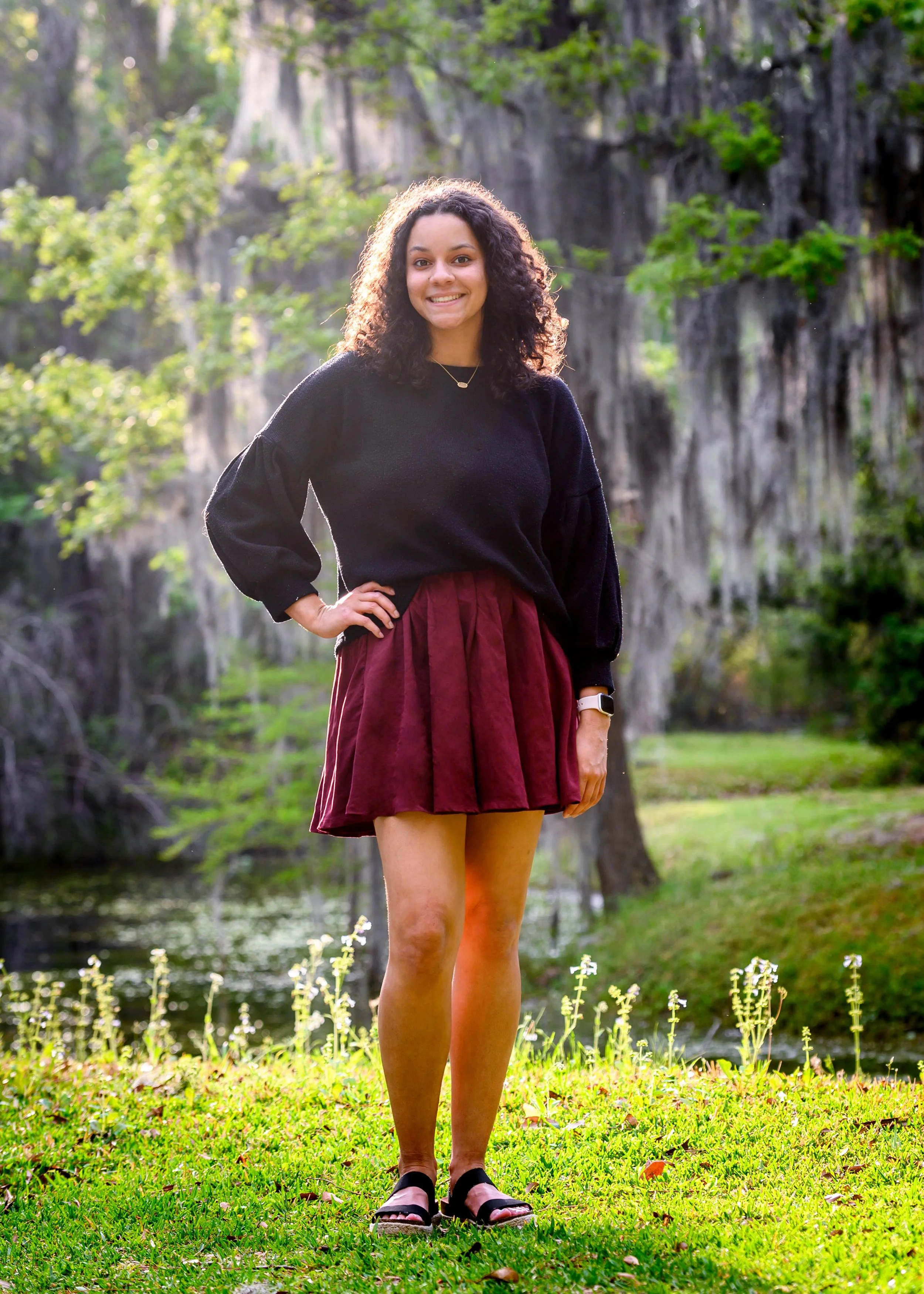A senior girl with curly hair smiling outdoors, standing on grass near a pond with trees in the background, wearing a black top, burgundy skirt, and sandals.