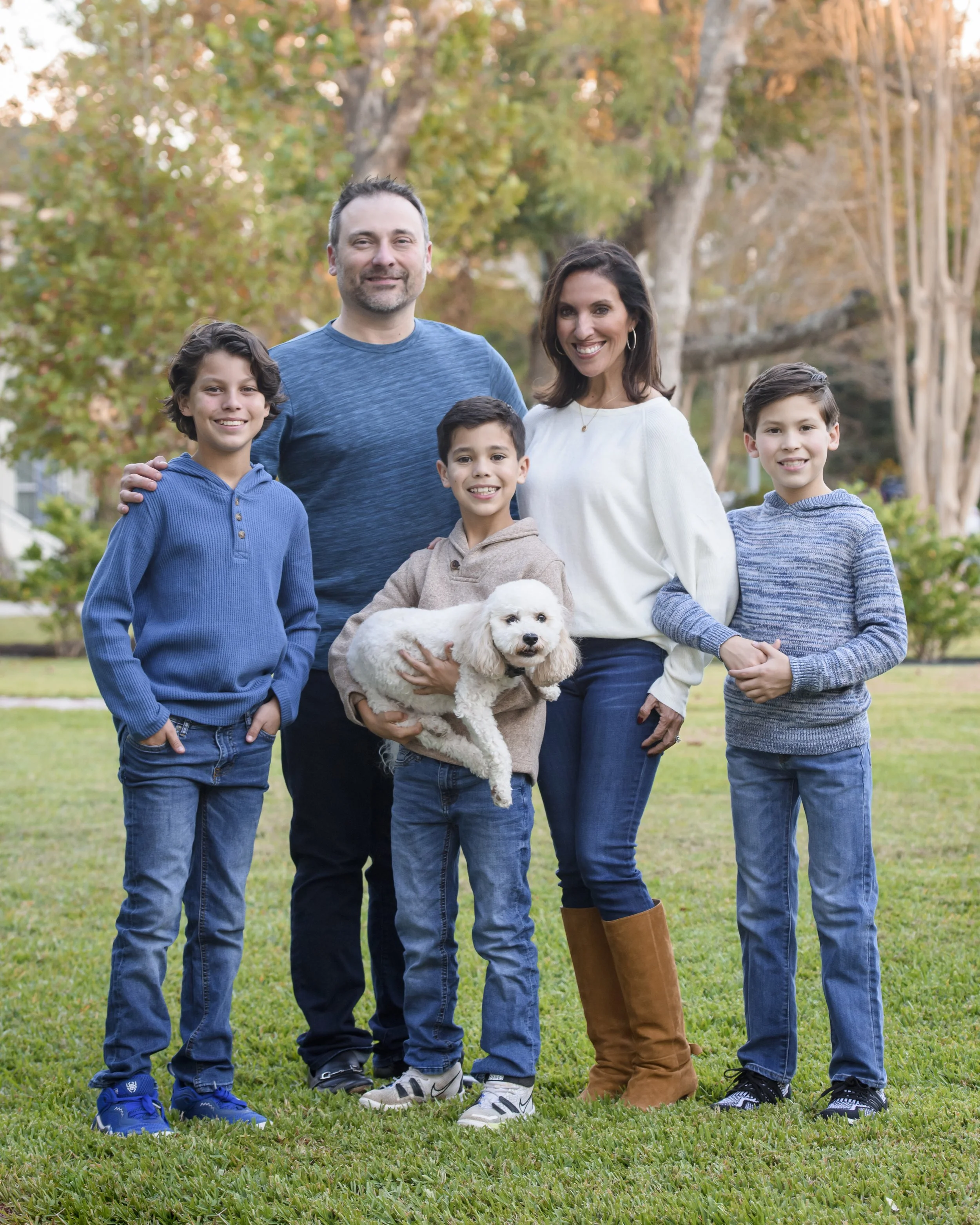 A family of five, including three children and two adults, standing outside on a grassy area in the fall, smiling at the camera with trees in the background. One child is holding a white dog.