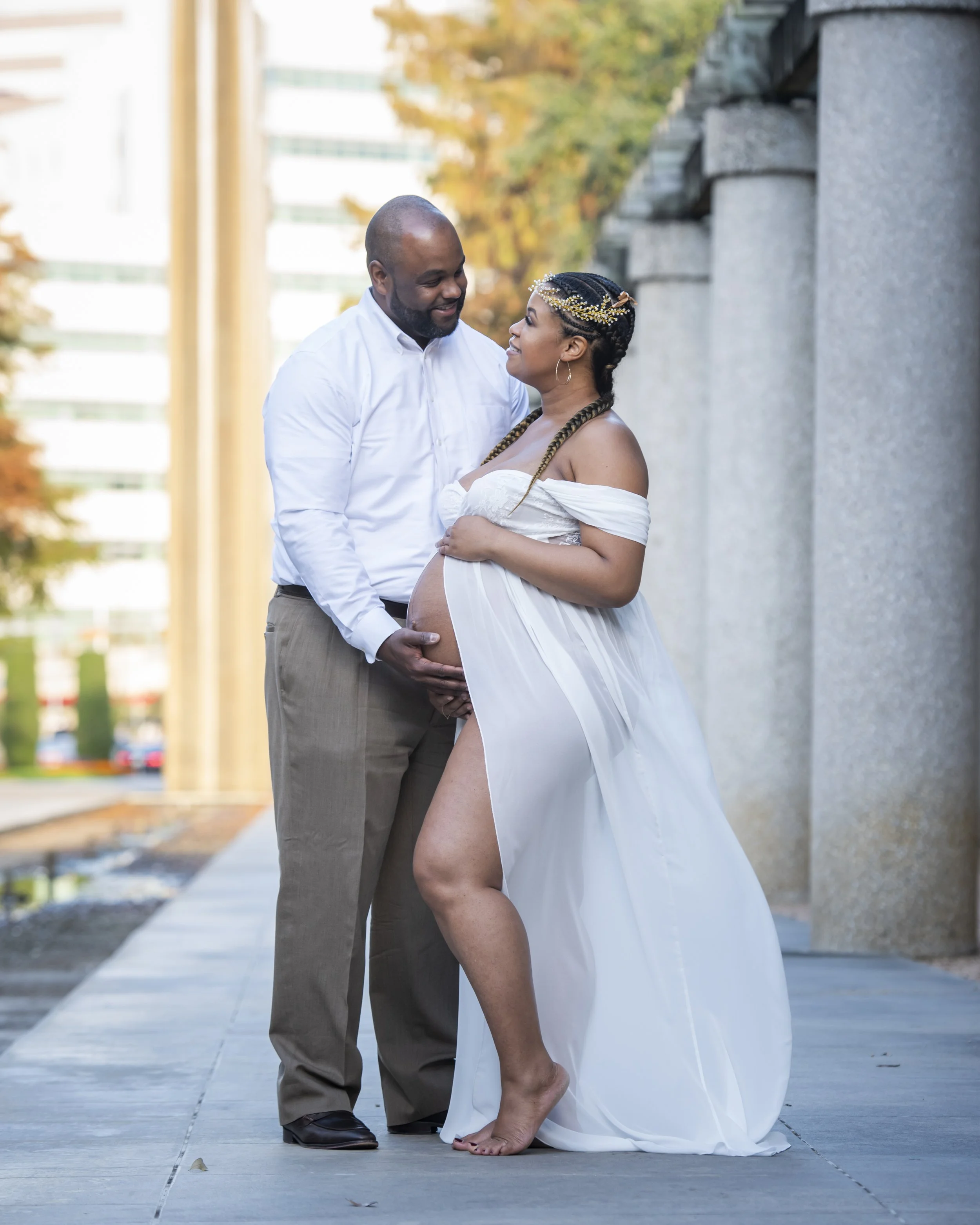 A pregnant woman in a white dress with off-shoulder sleeves and a thigh-high slit is standing on a sidewalk, looking at a smiling man in a white shirt and beige pants. The man is holding her belly, and she is wearing a headpiece with gold leaves and 