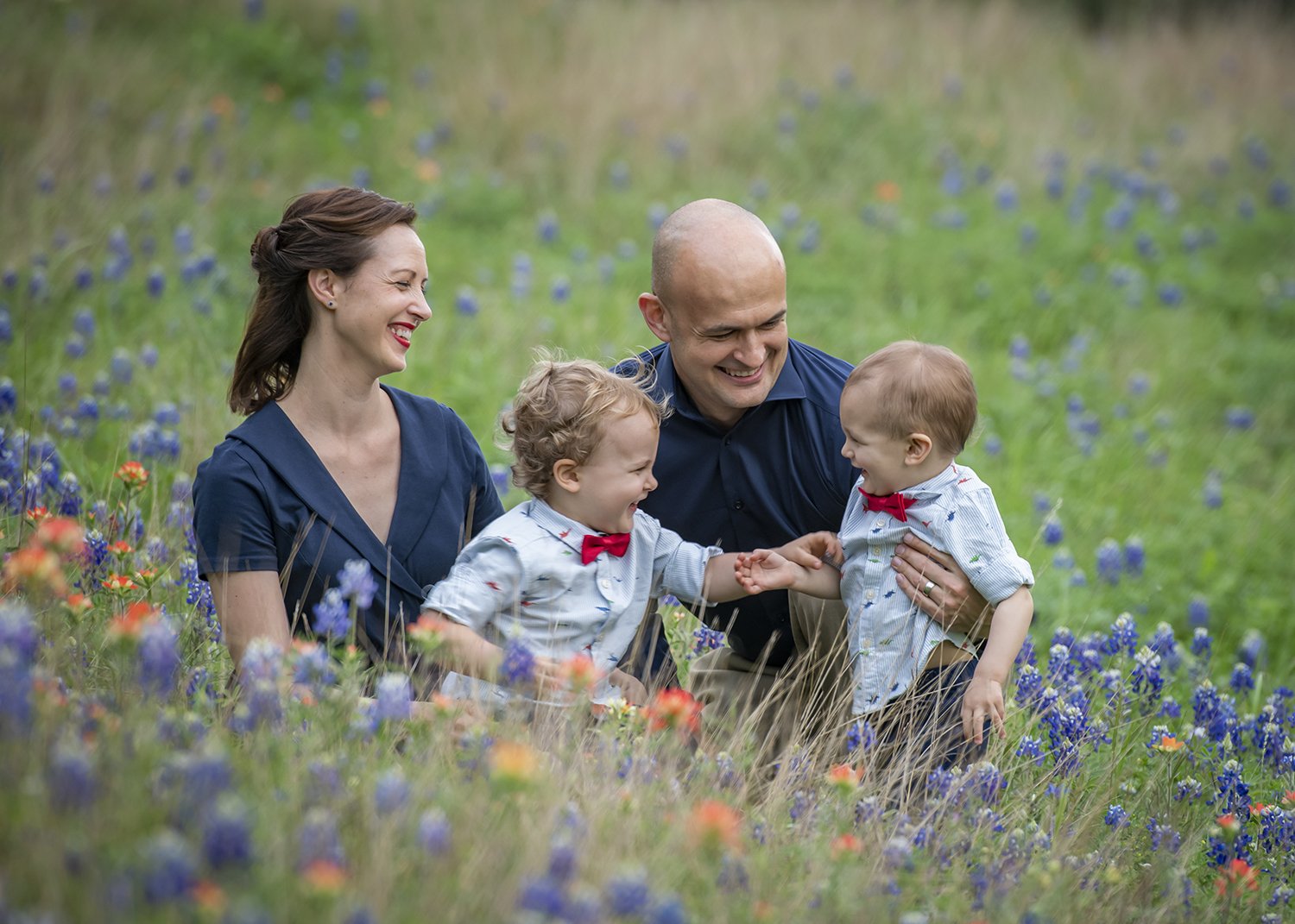 A happy family of four, including a mother, father, and two young boys, playing and laughing together in a colorful bluebonnets