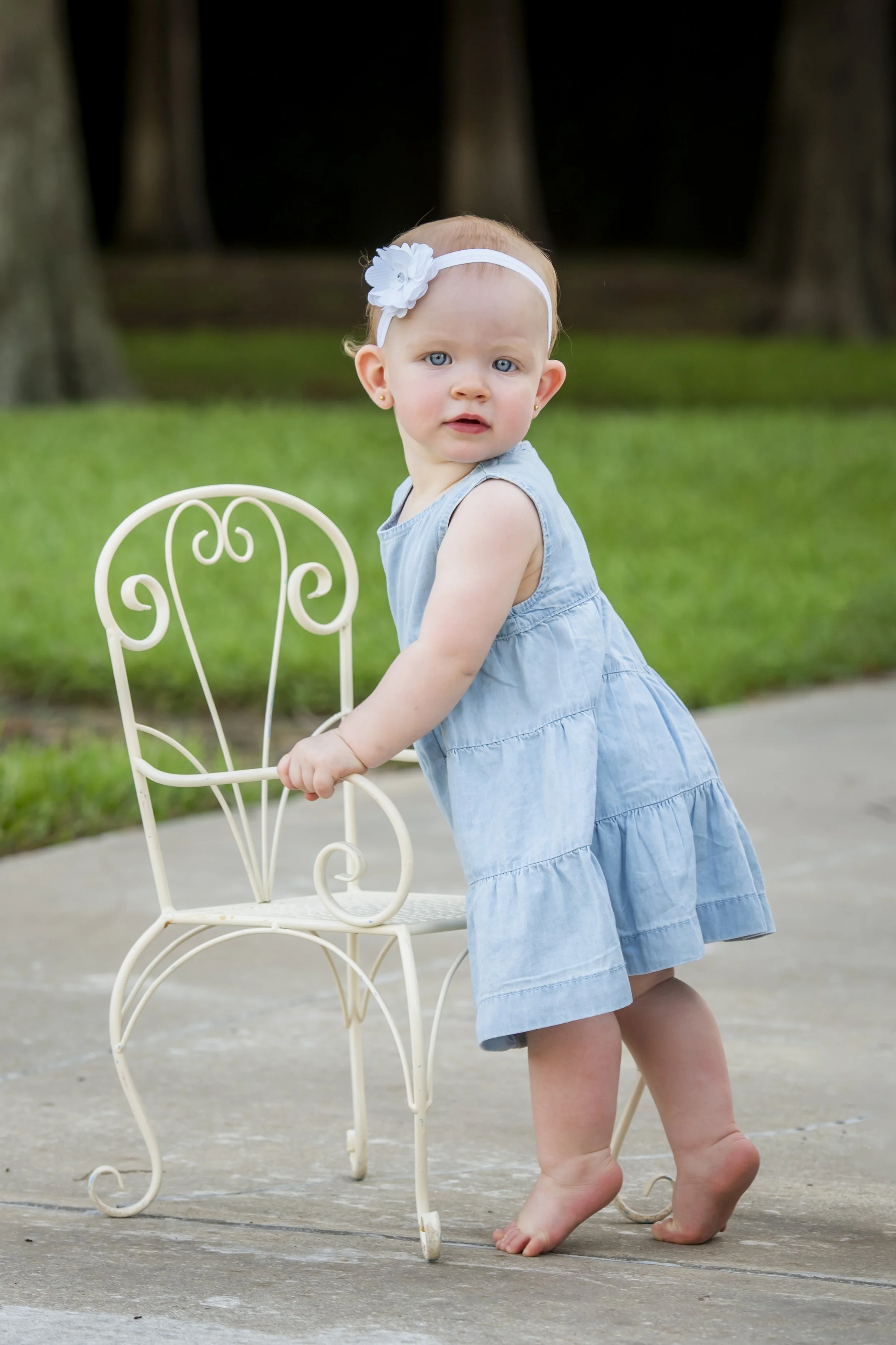 A young girl with blue eyes, wearing a light blue dress and a white headband with a flower, is standing outdoors on a concrete surface, holding onto a white decorative metal chair. She has small earrings and appears to be looking towards the camera w