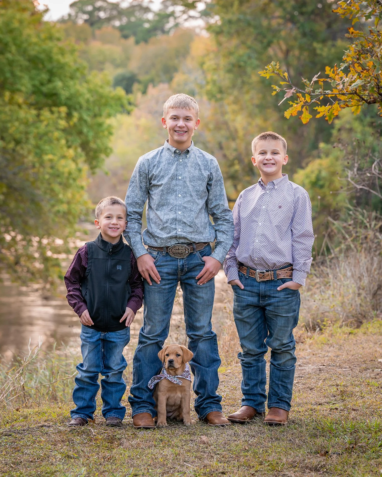 A family of three boys and a dog standing outdoors in a wooded area during fall with colorful leaves on the trees.
