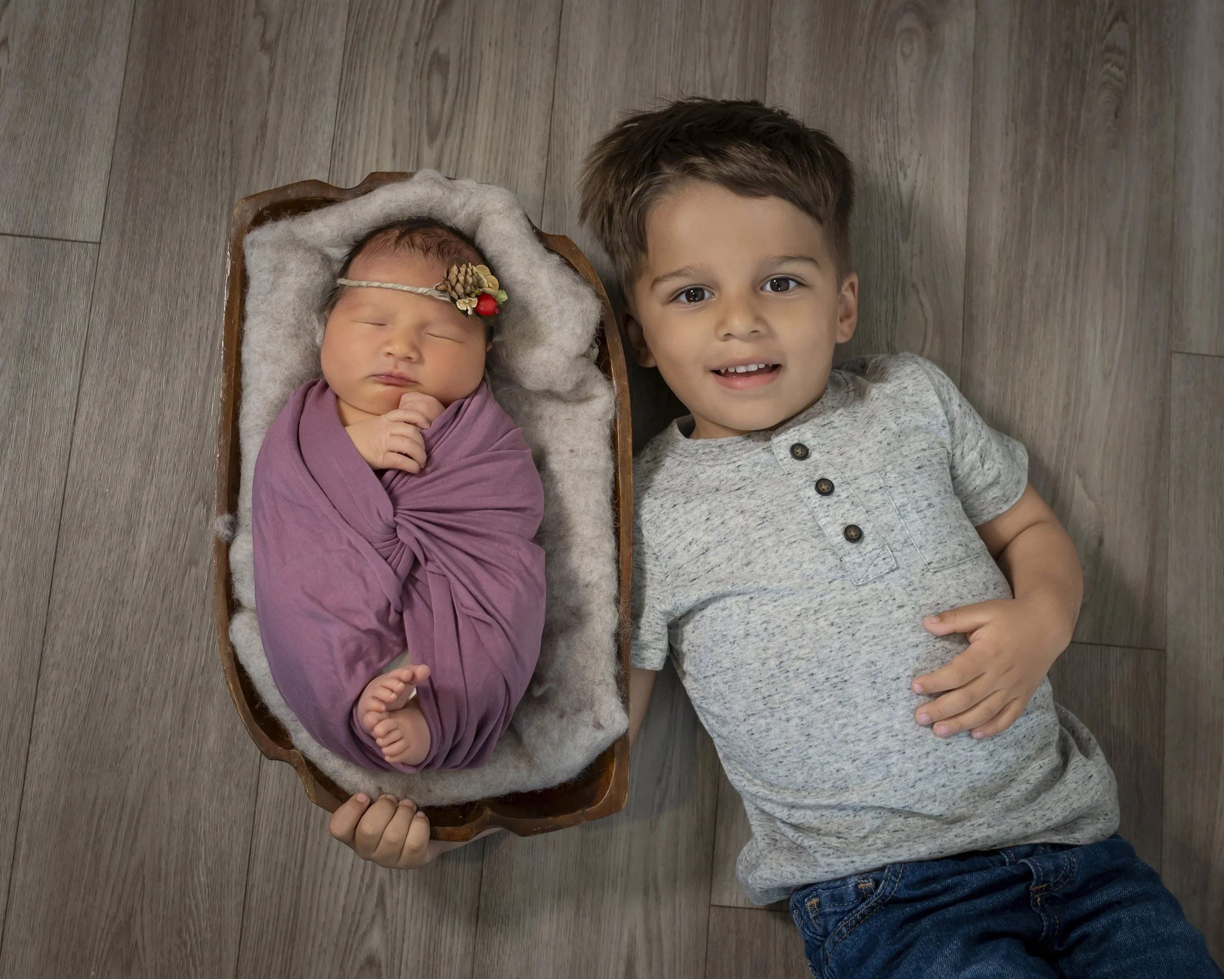 A young boy lying on the floor next to a sleeping newborn baby in a wooden cradle, both looking up at the camera.