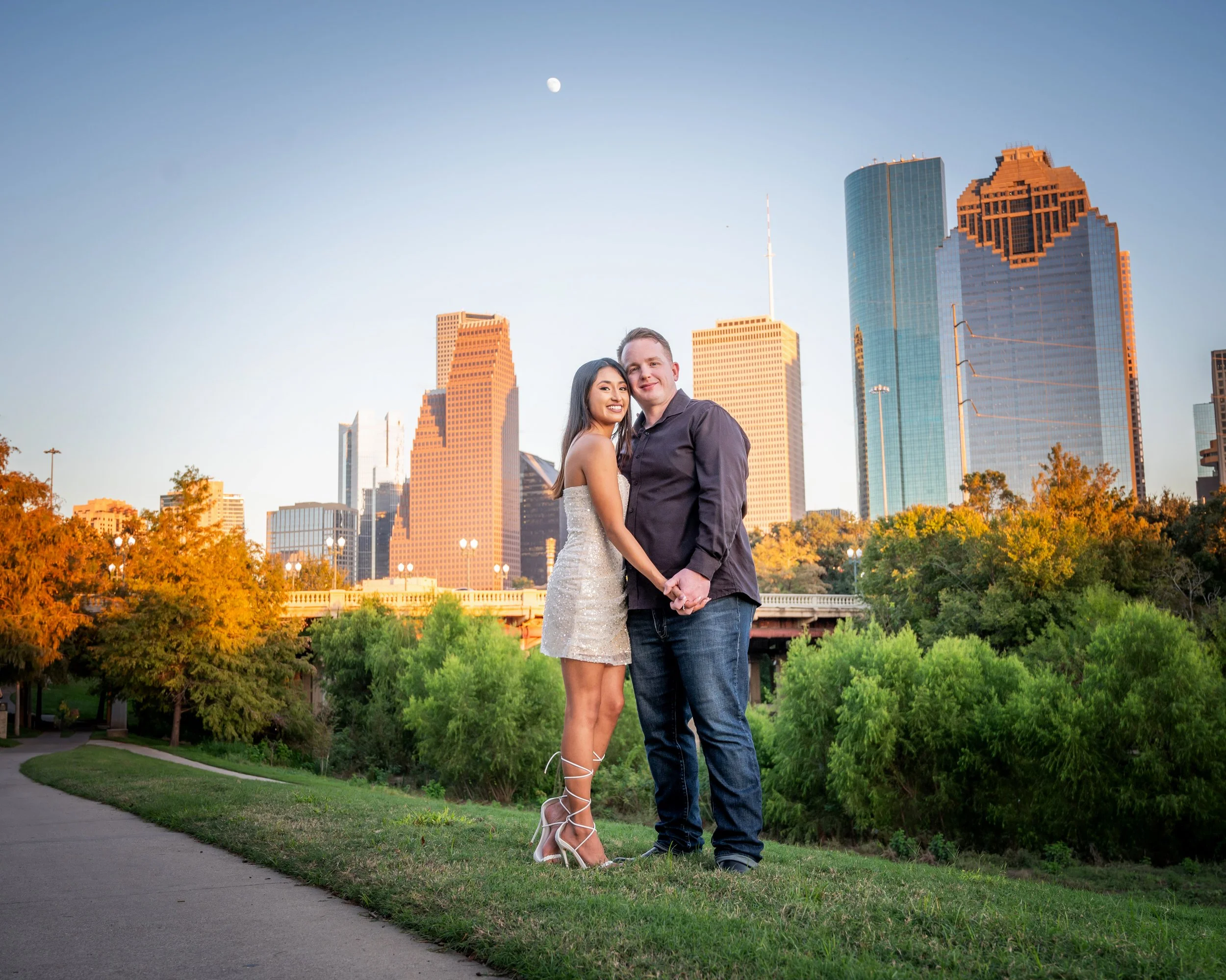 A couple standing close together outdoors in front of Houston skyline with tall modern buildings, trees with autumn foliage, on a clear evening with the moon visible in the sky.