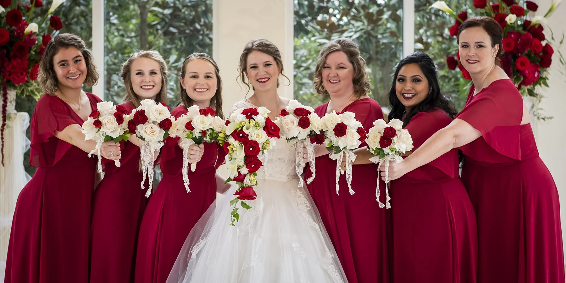 A bride and her bridesmaids standing indoors, holding bouquets of white and red roses. The women are dressed in matching red dresses, smiling, with a floral arrangement in the background.