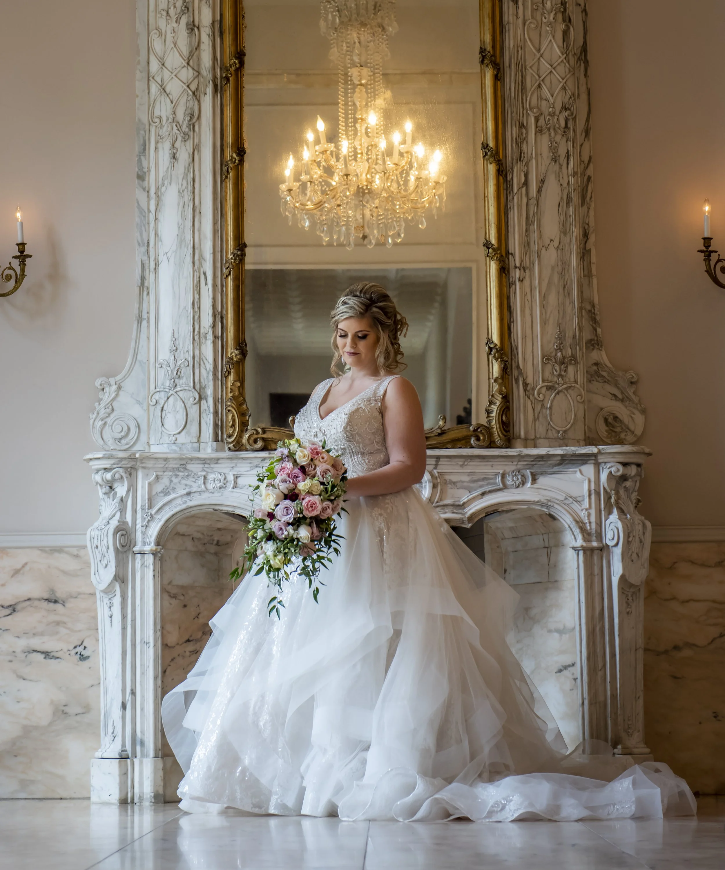 A woman in a wedding dress holding a bouquet of flowers, standing in an ornate room with a large mirror, a chandelier, and marble fireplace.