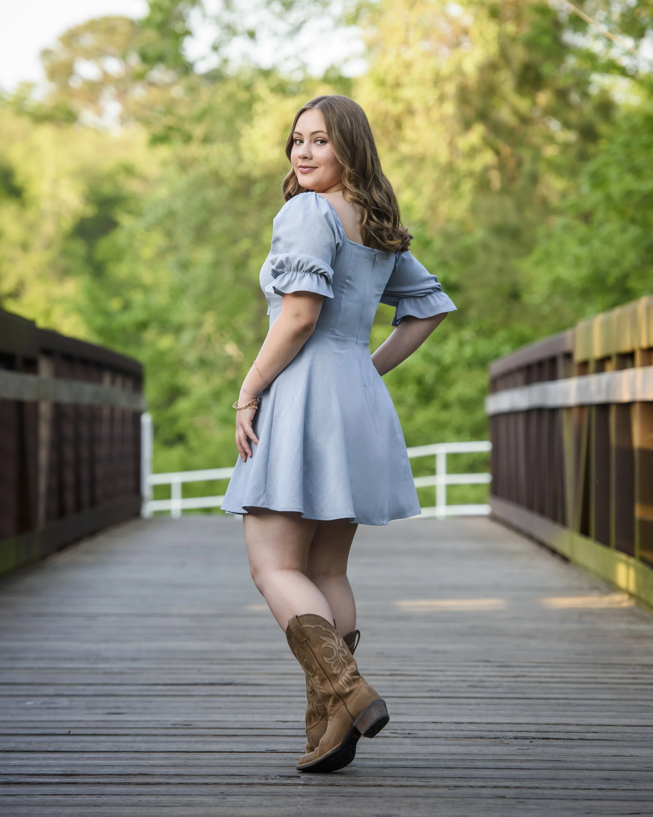 Senior girl in a blue dress and cowboy boots standing on a wooden bridge outdoors surrounded by greenery, posing with a slight smile and looking back.