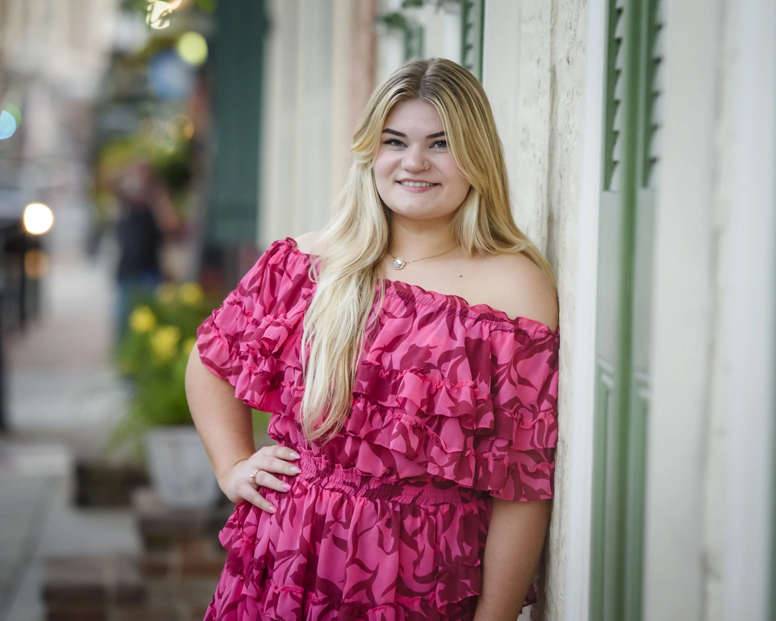 A senior girl with long blonde hair, wearing a pink ruffled off-shoulder dress, standing outdoors near a wall with her hand on her hip and smiling.