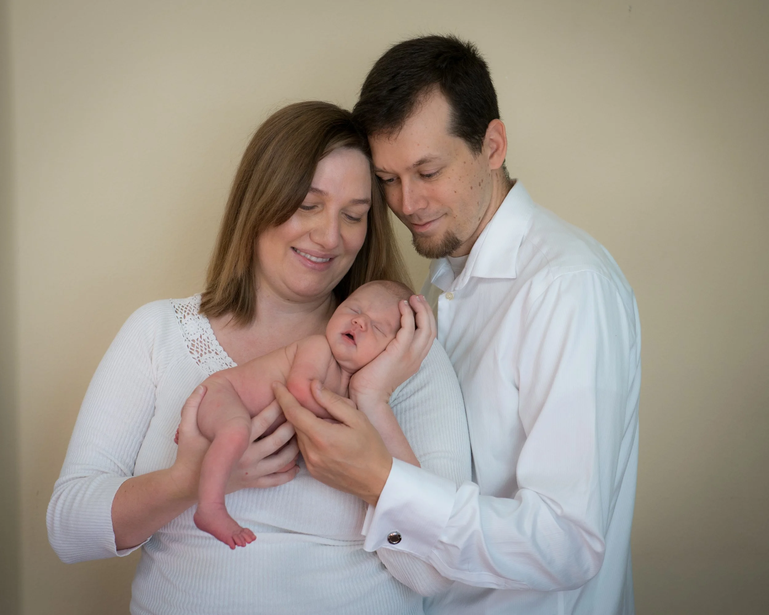 A woman and man holding a newborn baby, all smiling, against a plain beige background.
