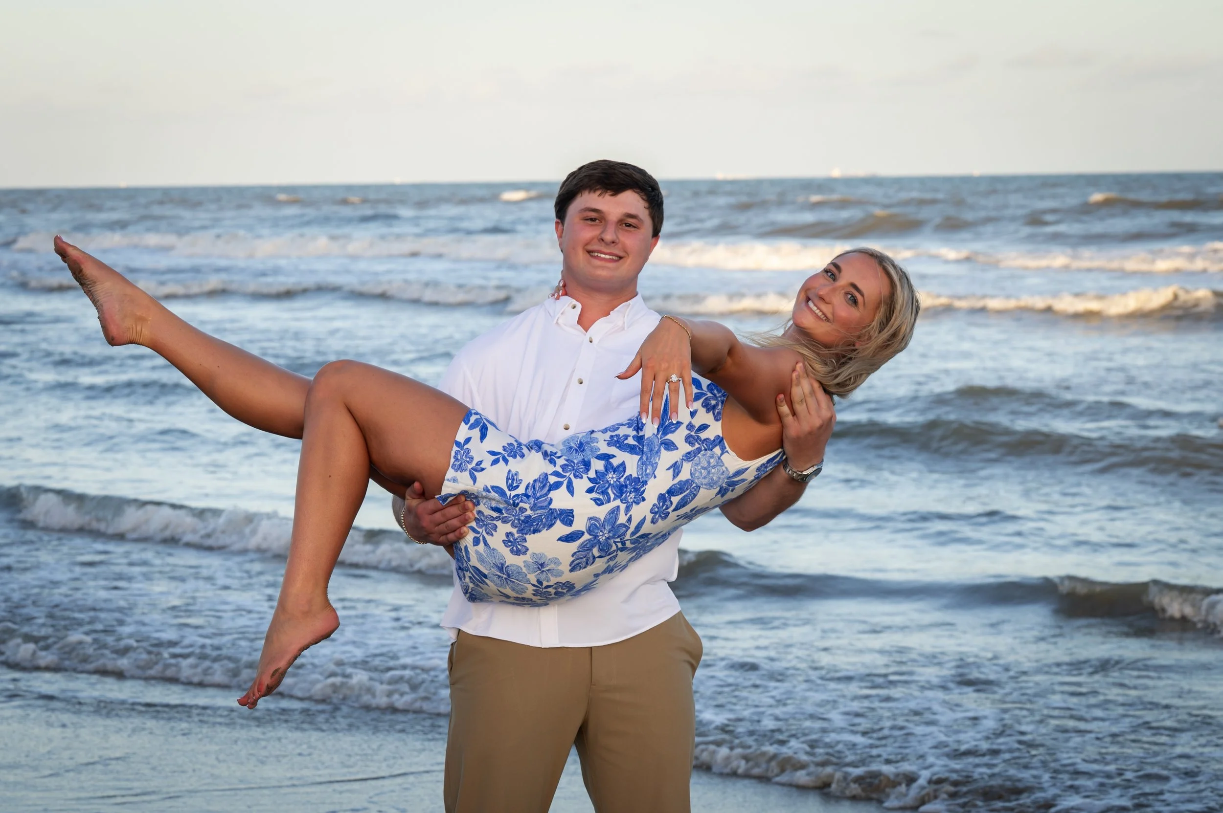 A man in a white shirt and tan pants is holding a smiling woman in a floral dress at the Galveston beach with waves in the background showing off engagement ring 