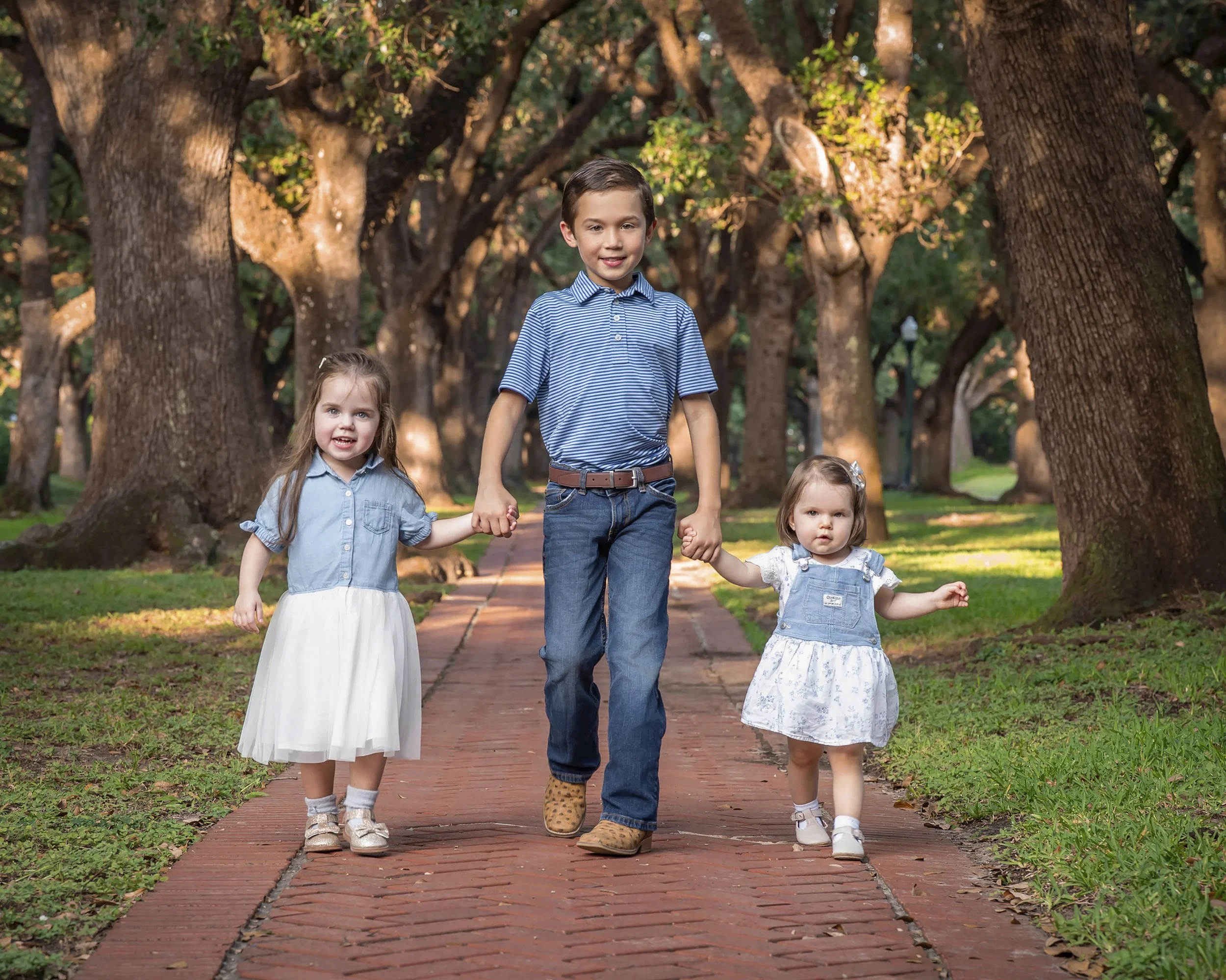 A boy and two girls holding hands while walking on a brick path through a park lined with large trees and green grass.