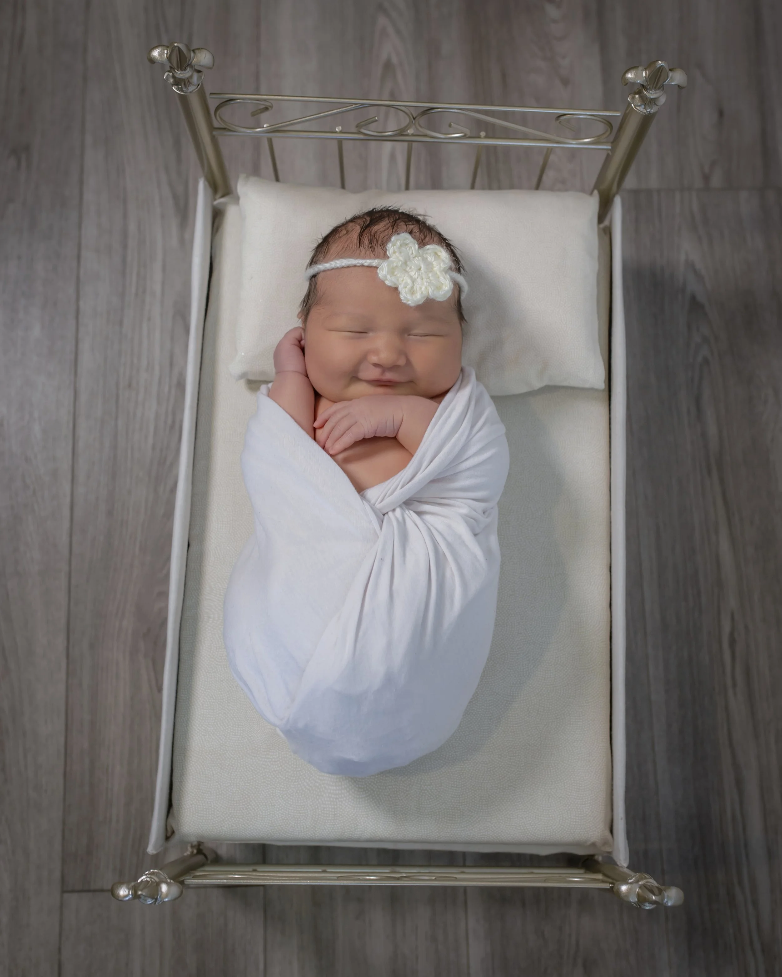 A smiling newborn baby wrapped in a white blanket and lying on a small white pillow in a miniature metal bed with a silver headboard and footboard, on a wooden floor.