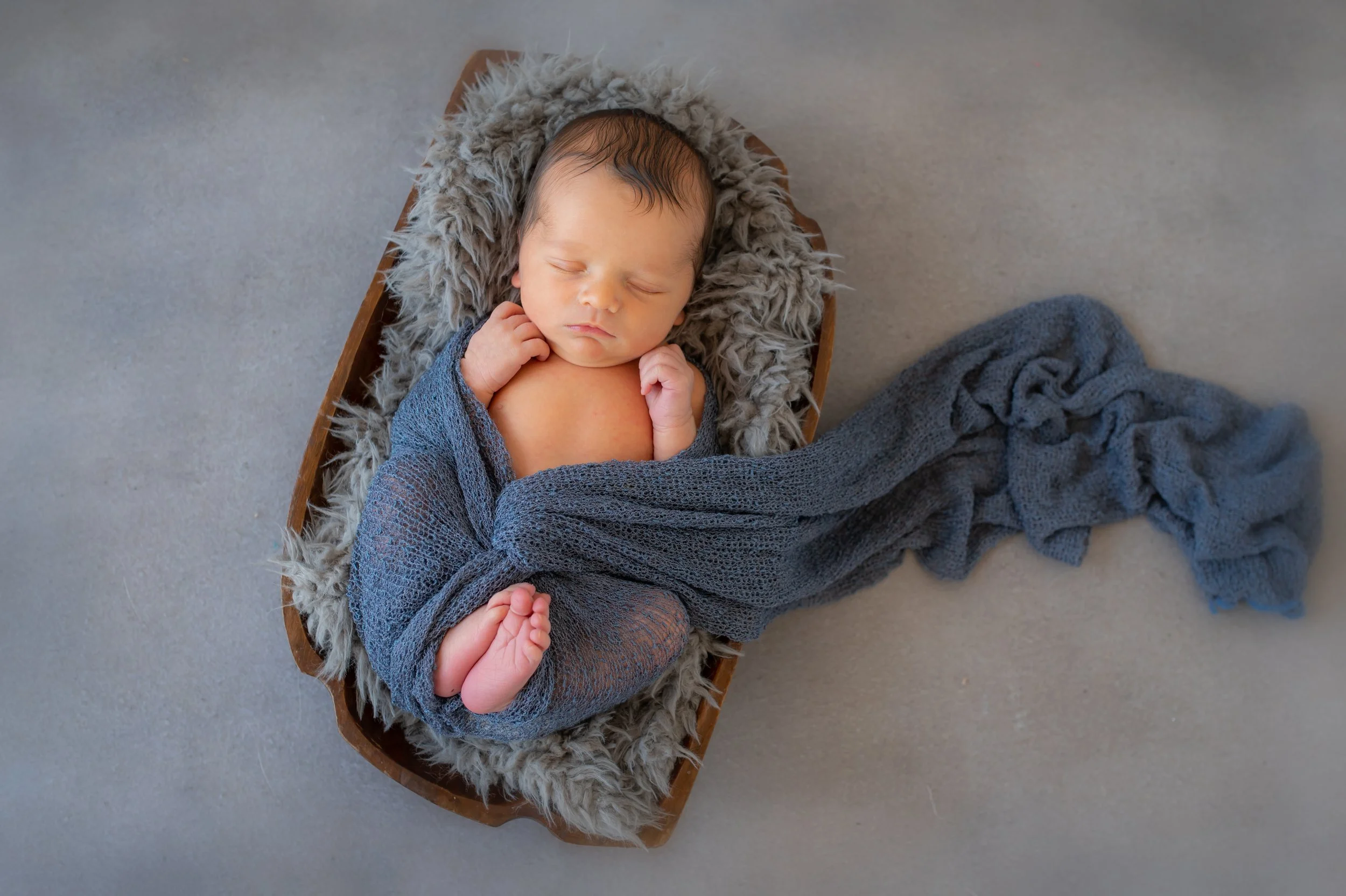 A sleeping newborn baby wrapped in a gray knitted blanket, lying in a wooden bowl lined with a furry gray material, on a gray floor.