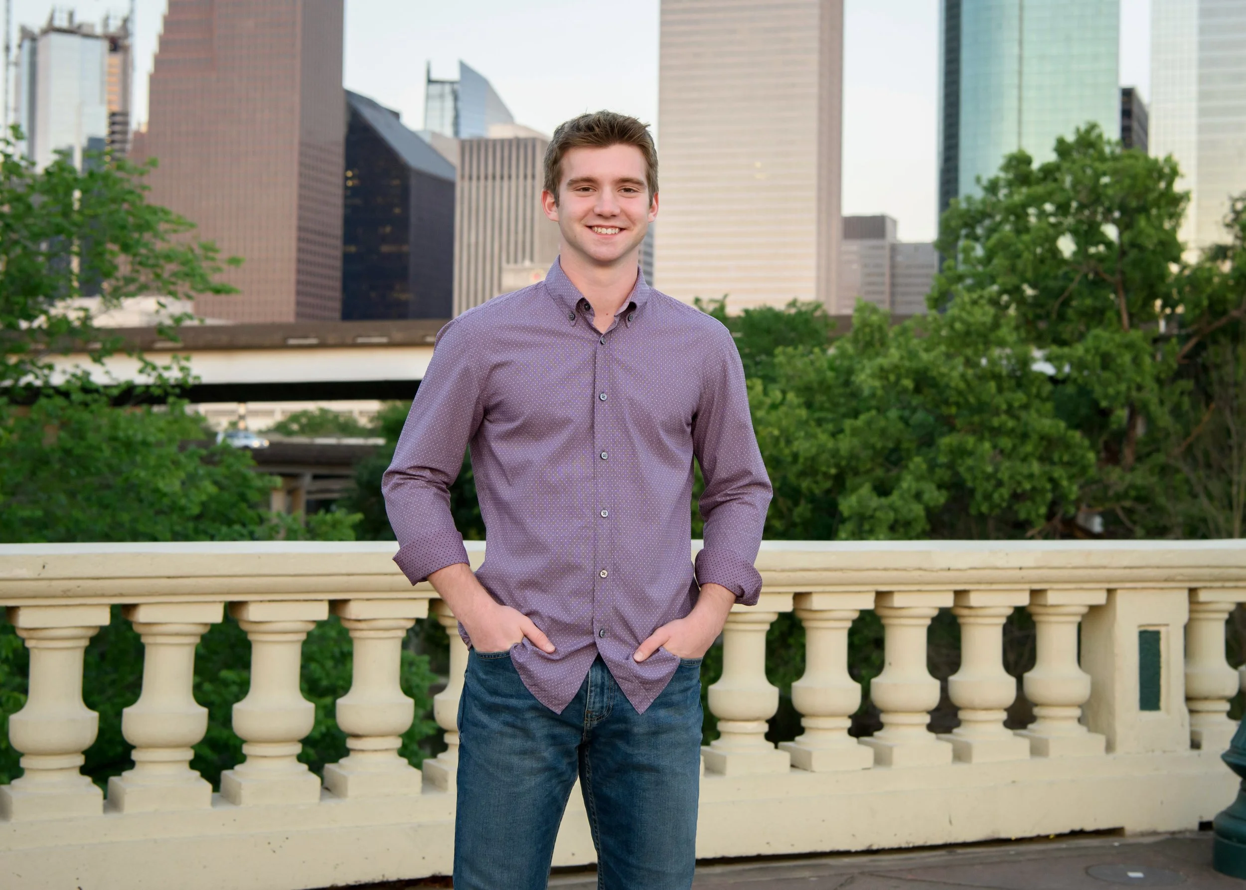 A senior guy in a purple button-up shirt standing outdoors near a white railing with green trees and modern skyscrapers in the background.