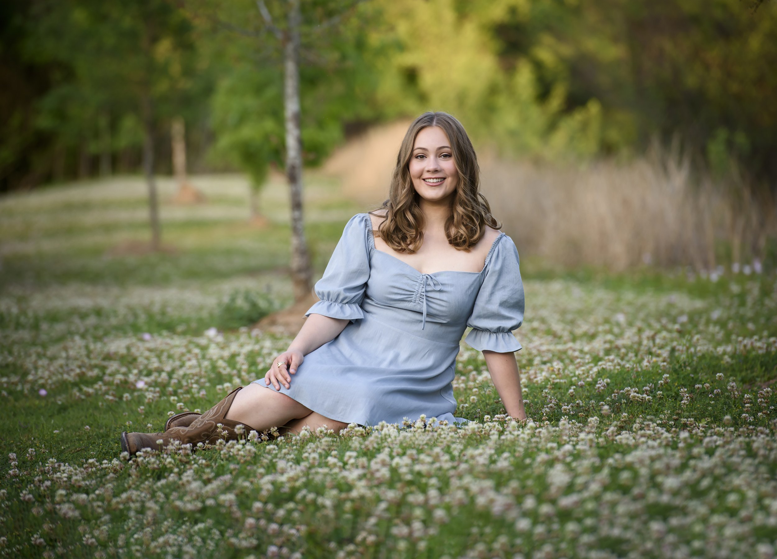 A senior girl with long brown curly hair, wearing a blue dress, smiling while sitting on a grassy field with small white flowers, in a park with trees in the background.