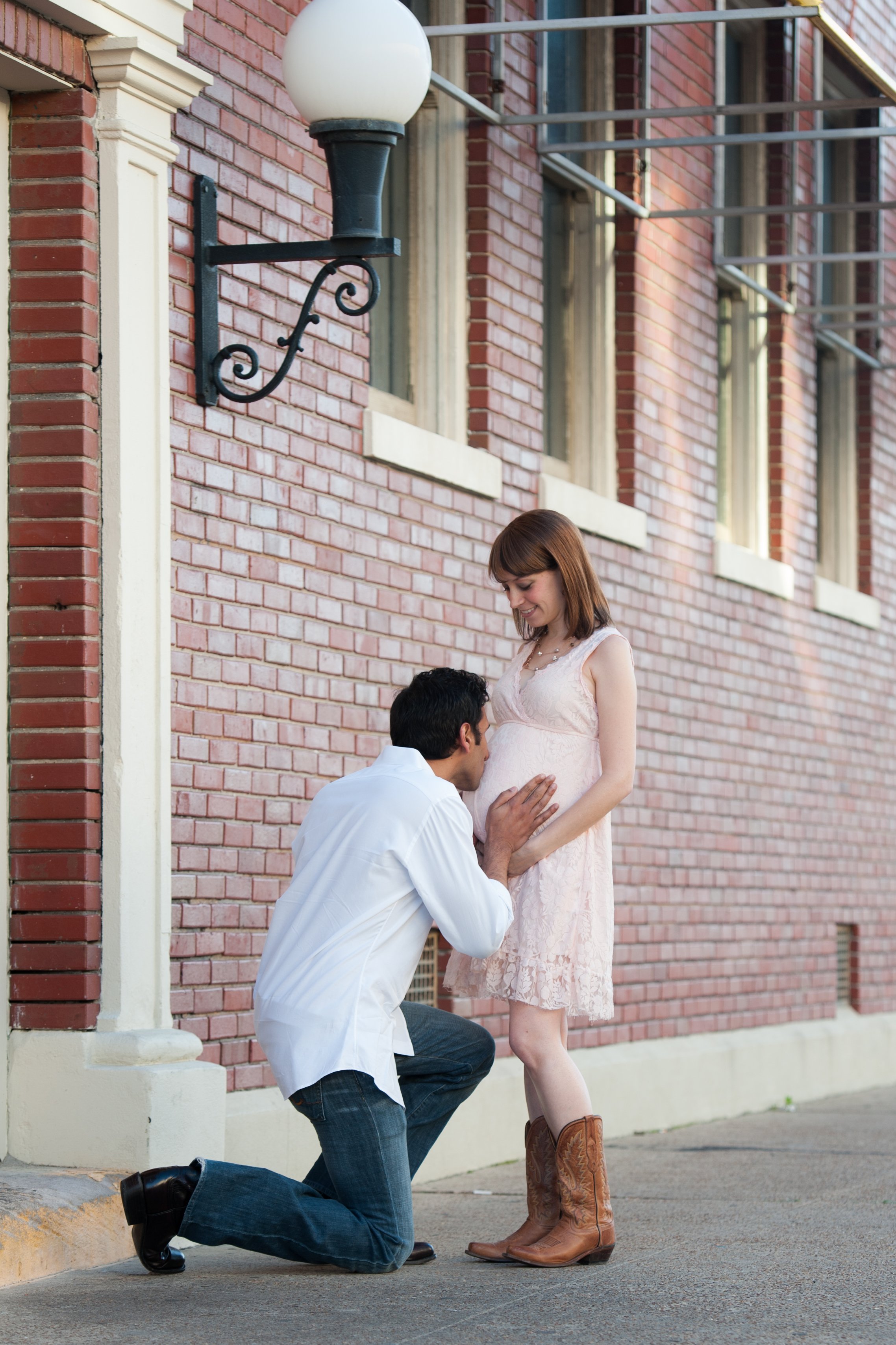 A man kneeling with one knee on the ground proposing to a pregnant woman standing in front of a brick building. The woman is smiling and wearing a light pink dress and brown cowboy boots. The man is in a white shirt and dark jeans.