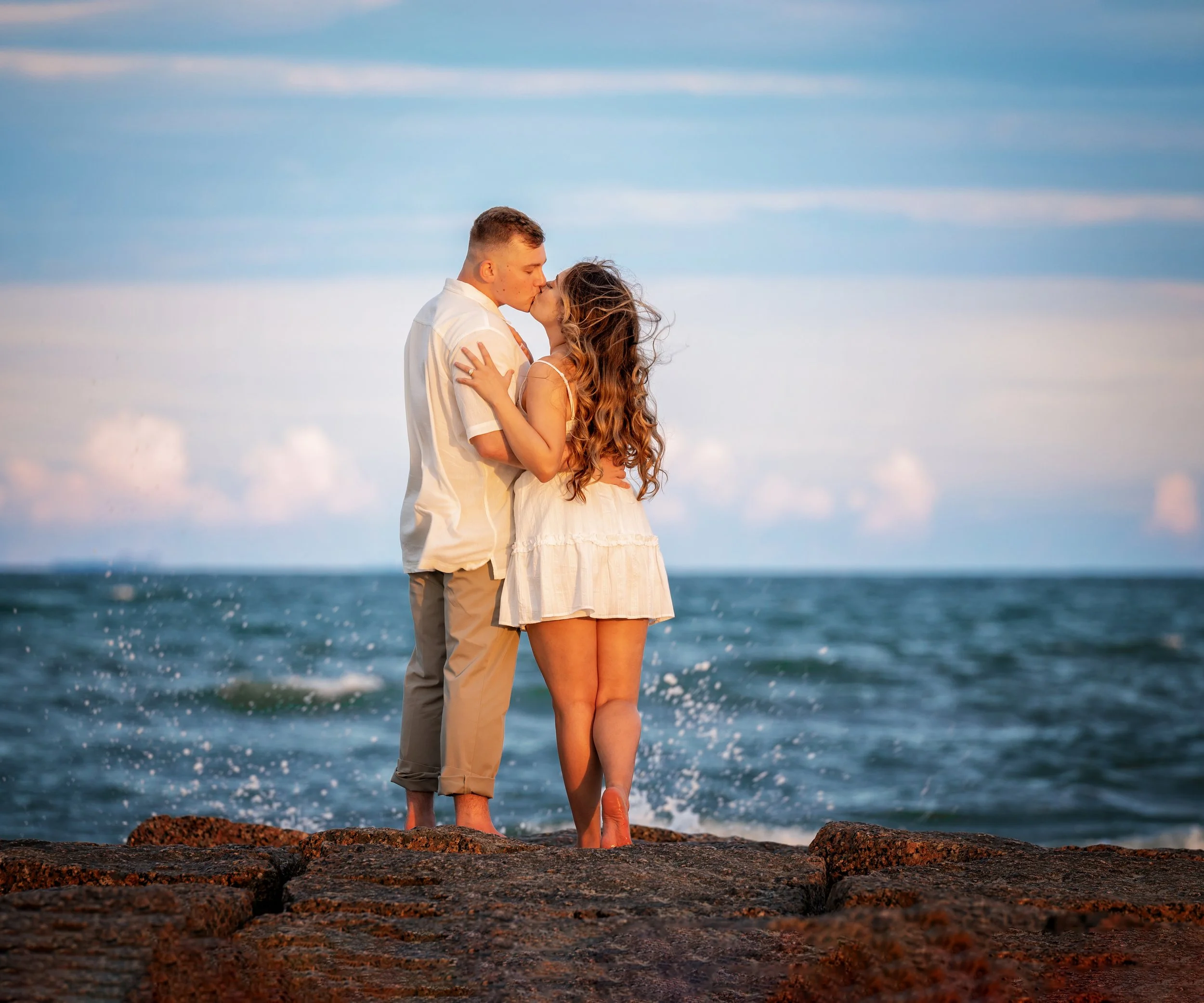 A young couple kissing and holding each other on a rocky beach, with the ocean and a clear sky in the background during sunset.