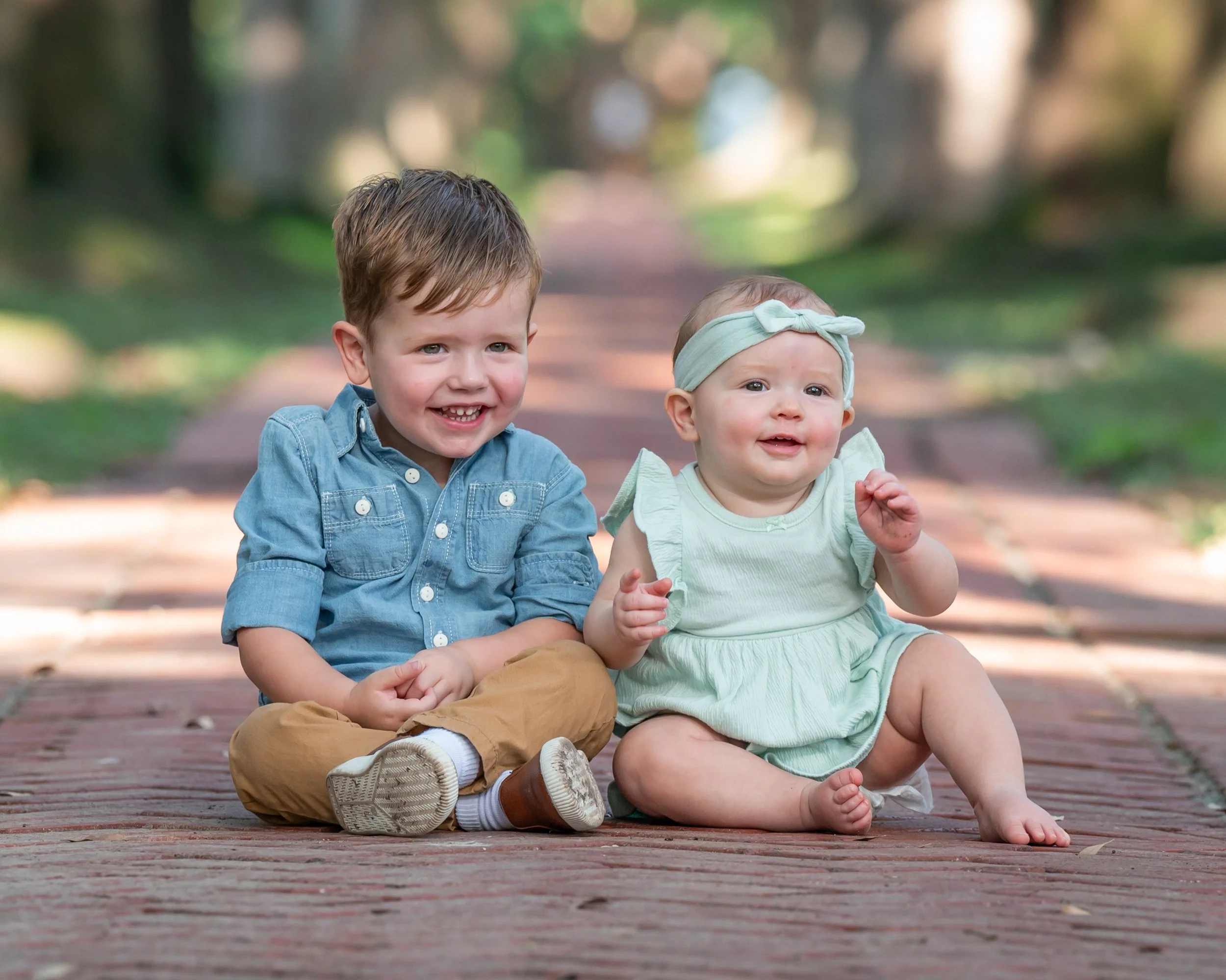 Two young children sitting on a wooden pathway in a park, smiling and playing with nature around them.
