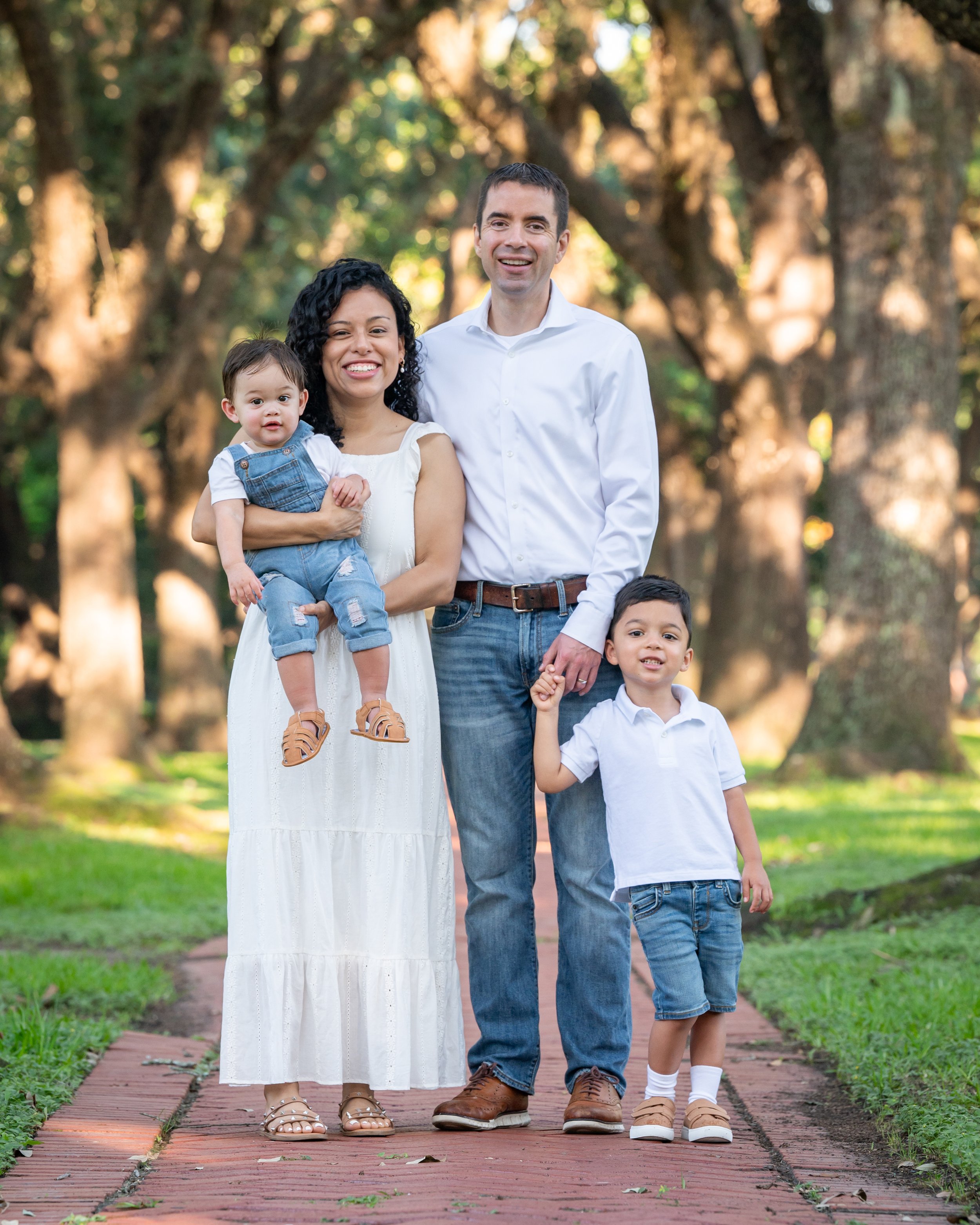 A family of five walking on a brick path in a park, with trees in the background. The mother is holding a toddler, and they are all smiling. The father and two older children are walking alongside them.