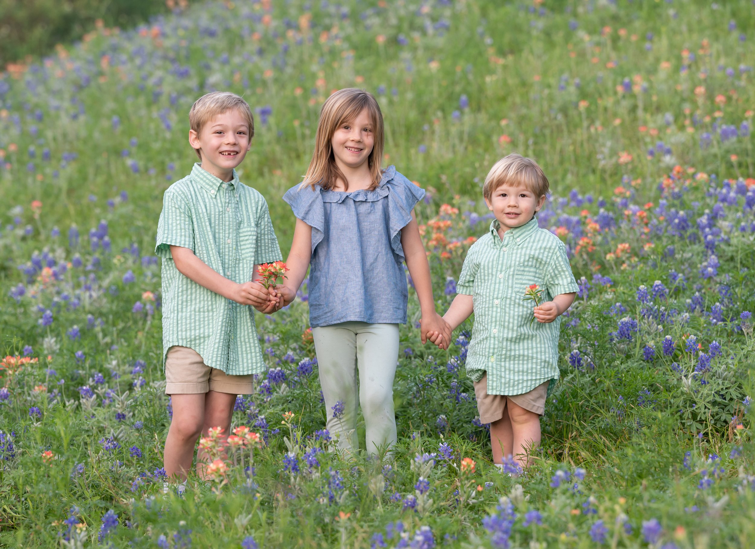 Three children, two boys and one girl, holding hands and flowers in a field of bluebonnets and orange flowers, smiling at the camera.