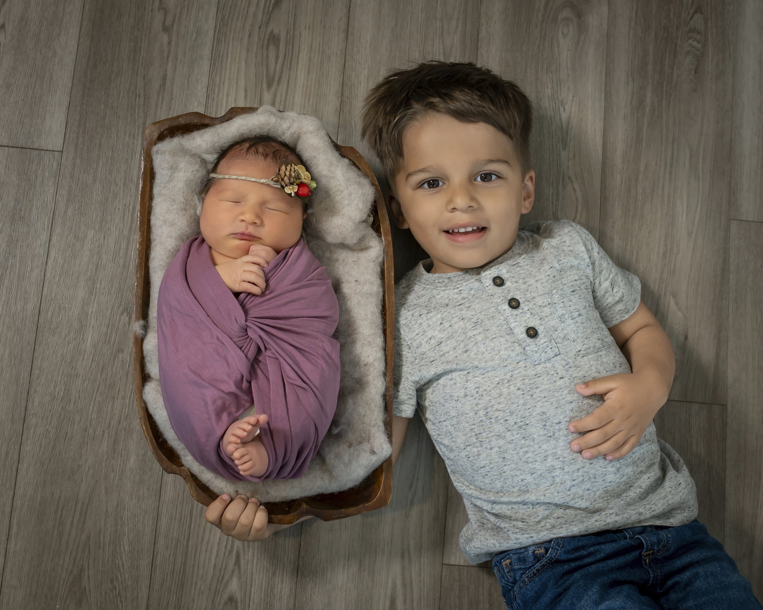 A young boy and a newborn baby laying on a wooden floor, with the boy smiling at the camera. The baby is wrapped in a pink blanket and wears a floral headband, sleeping in a small wooden cradle.