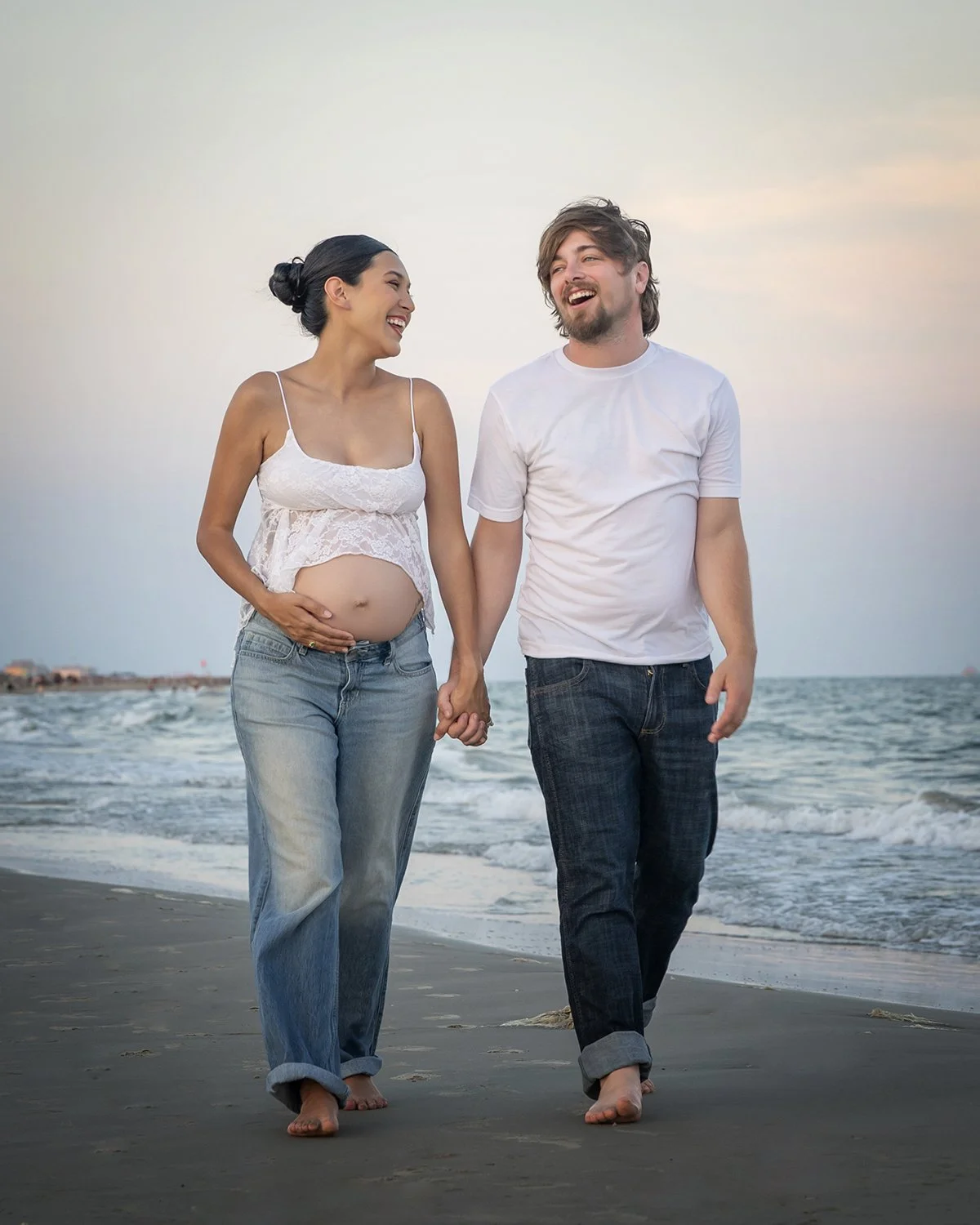 A pregnant woman and a man walking hand in hand on Galveston beach, smiling and looking at each other, with the ocean and sky in the background.