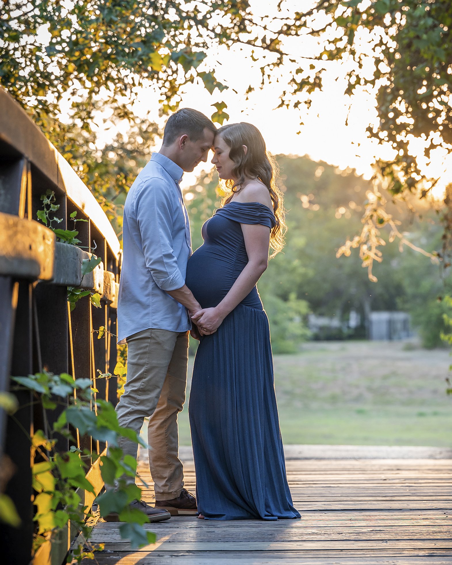 A man and a pregnant woman standing close together outdoors on a wooden deck, touching foreheads with eyes closed, illuminated by warm sunset light, surrounded by trees and greenery.