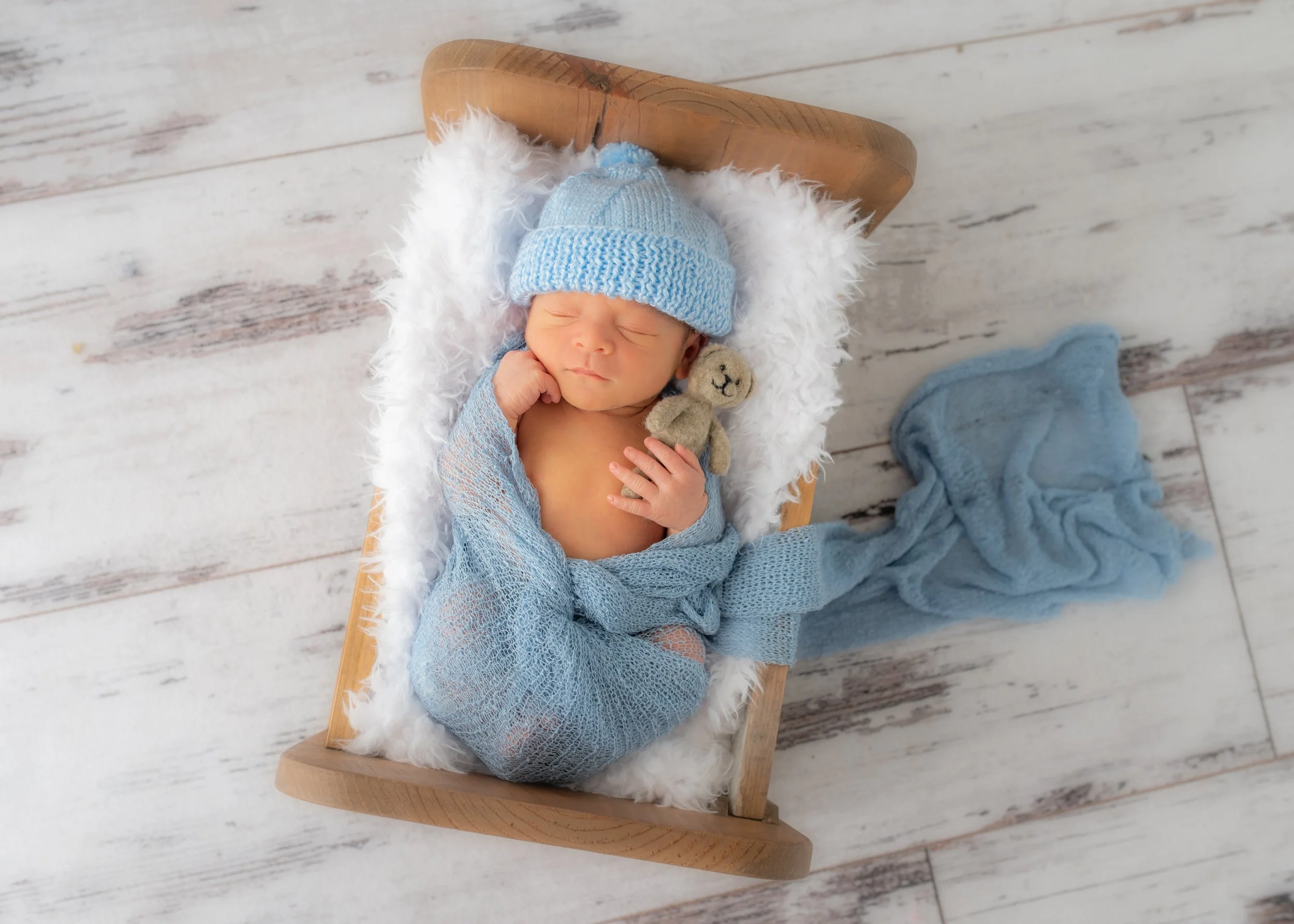 A newborn baby dressed in a blue knit hat and a matching blue blanket, lying on a fluffy white blanket in a small wooden bed with a furry liner, on a white wooden floor with a stuffed bear and a blue blanket nearby.