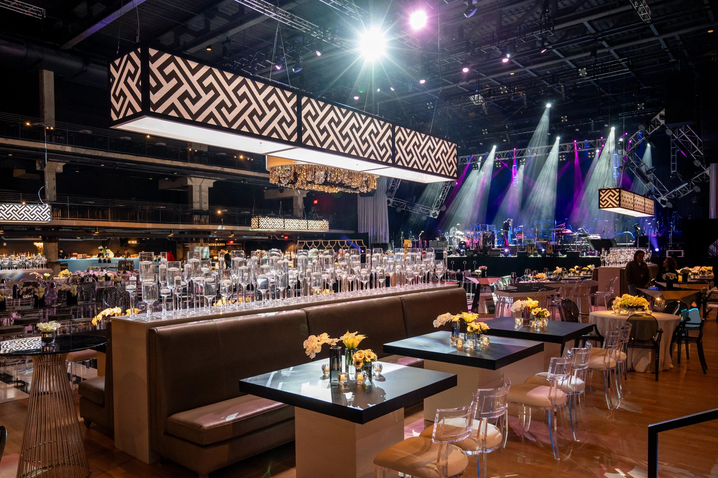 A large indoor event space with a stage, decorative lighting, and hanging screens displaying geometric patterns. There are tables with floral centerpieces and glassware, and concert equipment on the stage.