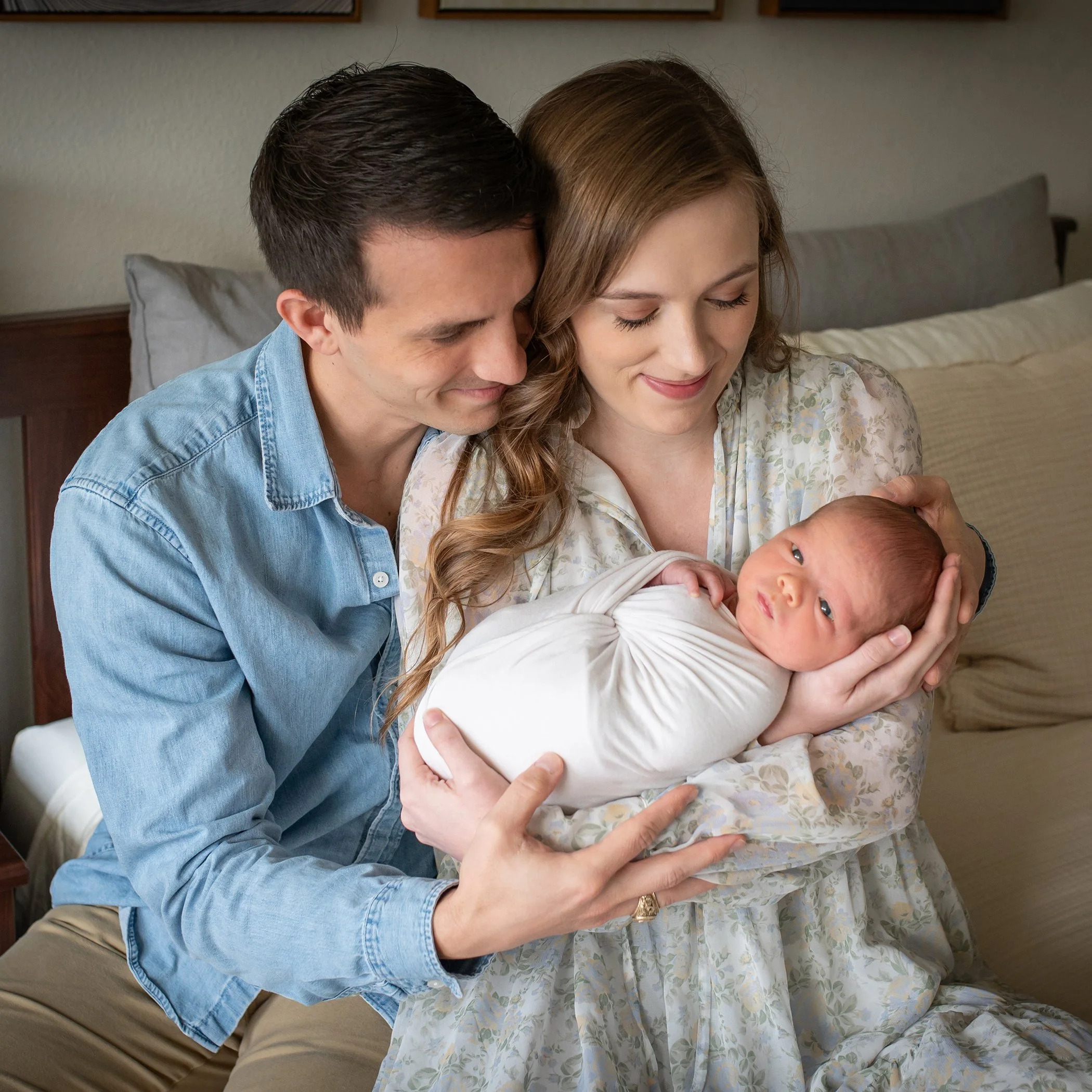A man and woman holding a newborn baby. The man is resting his forehead against the woman's temple, both smiling gently, in a cozy indoor setting.